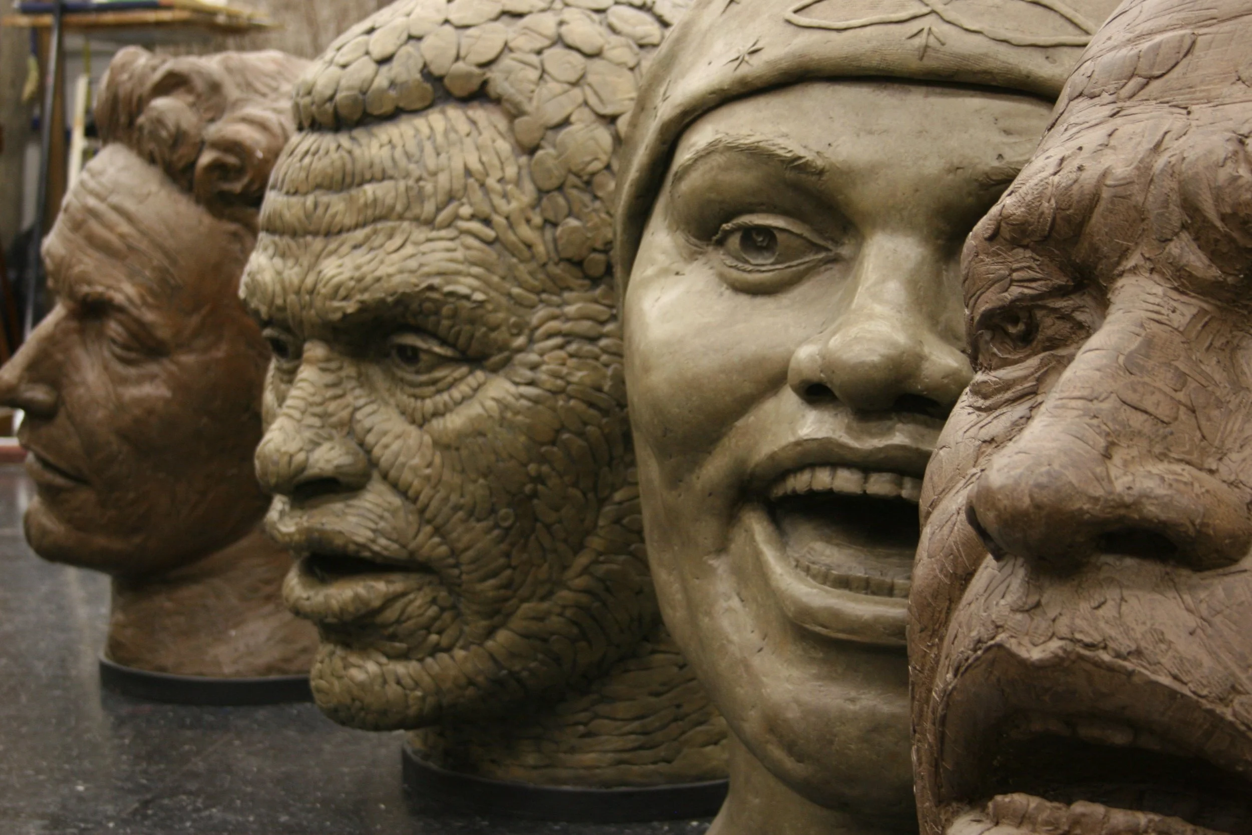 Close-up of multiple detailed sculptures of human faces of plaster painted different colors, arranged in a row. Cultural Heroes