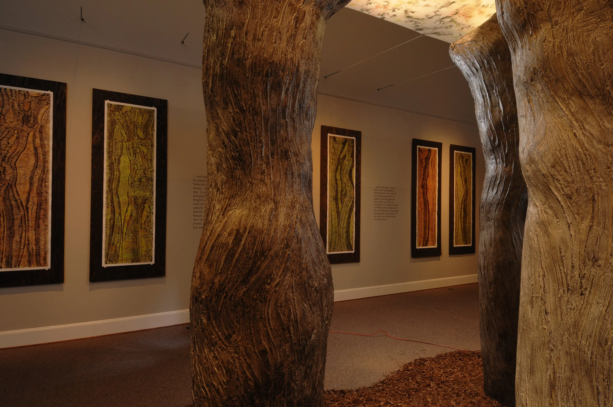 Art exhibit featuring massive tree trunk sculptures inside a gallery with framed abstract artwork on the wall. Dream Forest installation. 