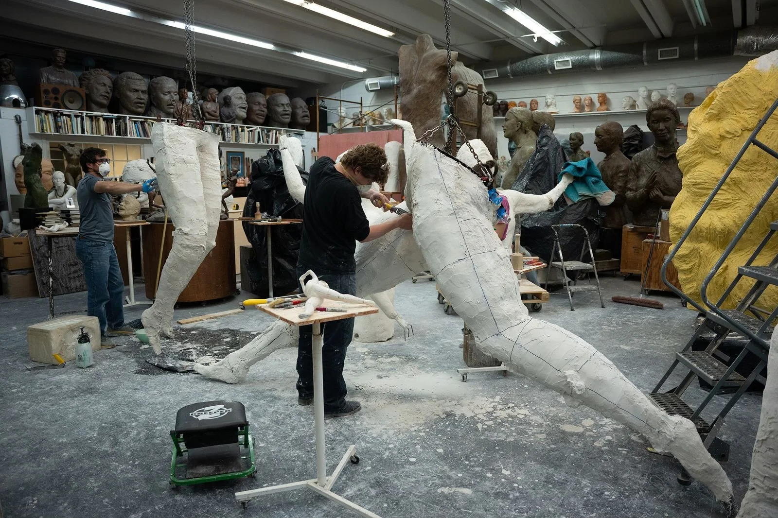 People working on creating a large sculpture in an art studio filled with busts, sculptures, and art supplies.