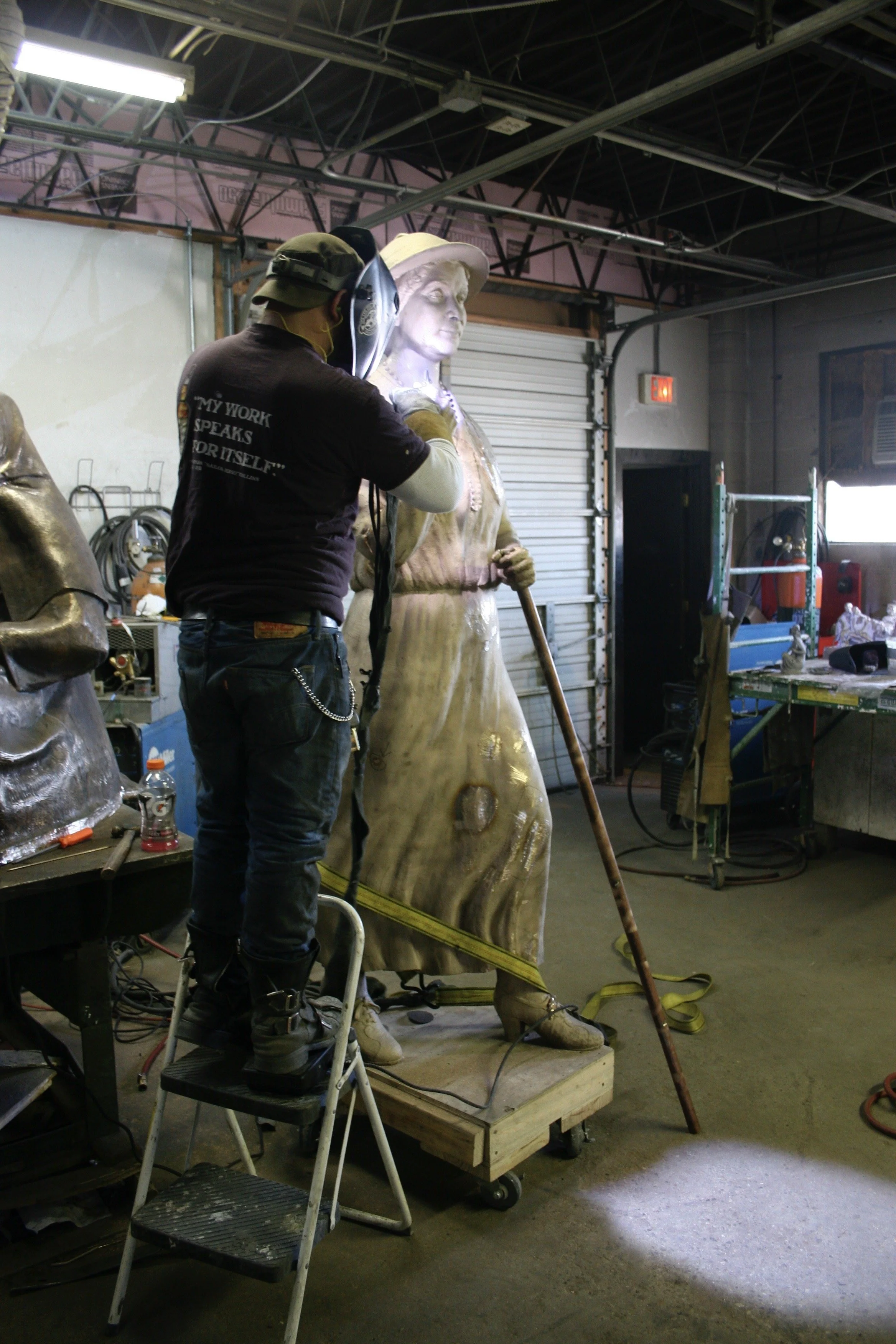 A person is working in a workshop, chasing a bronze sculpture of a woman wearing a hat, standing on a wooden base. The workshop contains tools and equipment, with exposed ceiling pipes and a garage door in the background.