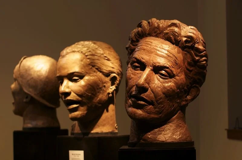 Three sculpted busts of people's heads displayed on black pedestals in an art gallery.  Cultural Heroes