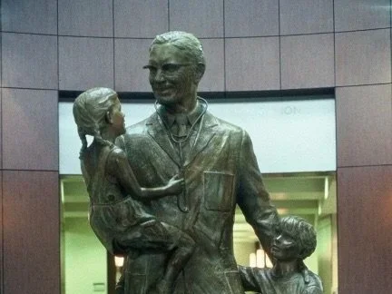 Bronze sculpture of a father in a military uniform holding his daughter and standing with his son, in an indoor setting.