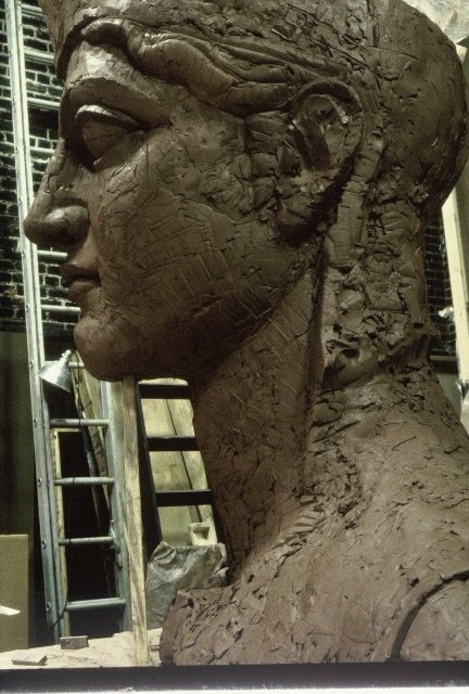 Close-up of a large clay sculpture of a woman's profile with detailed facial features and hairstyle, in an artist's studio with scaffolding and tools in the background. Athena Parthenos in Parthenon Museum Nashville. 