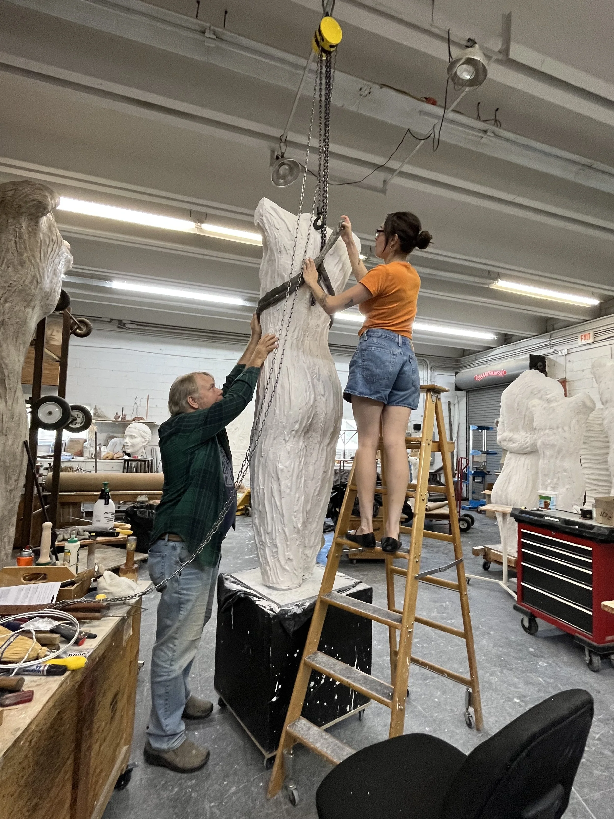 Two people working on a large, white sculpture of a figure in an art studio. One person is on a ladder adjusting the sculpture, while the other is holding it steady. The studio has various art supplies and tools around.