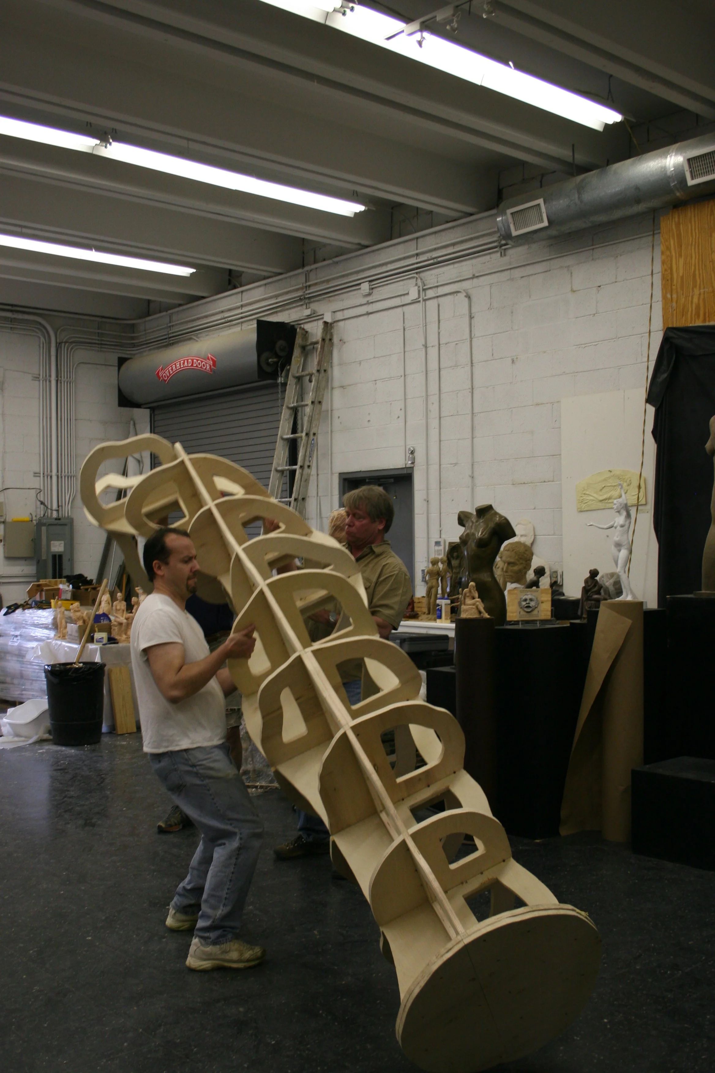 Two people are working together to build a large wooden sculpture that resembles a staircase or coiled structure in a workshop space. The background shows various art sculptures and supplies.