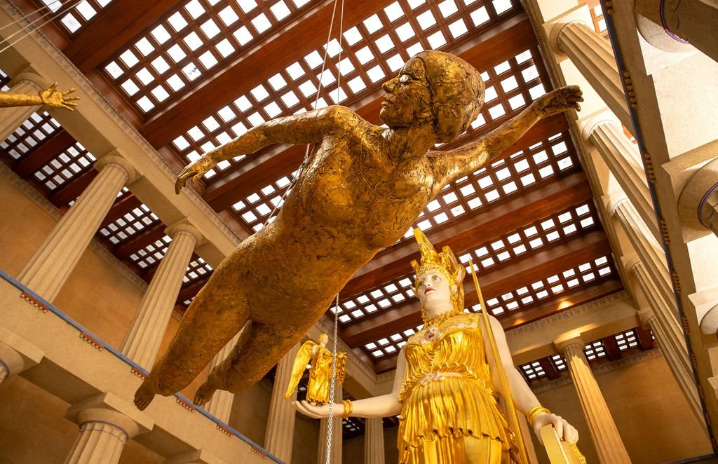 A large golden statue of a woman in a dress and crown holding a small statue of an angel, with a suspended bronze figure of a person reaching out, inside a building with columns and a checkered wooden ceiling.