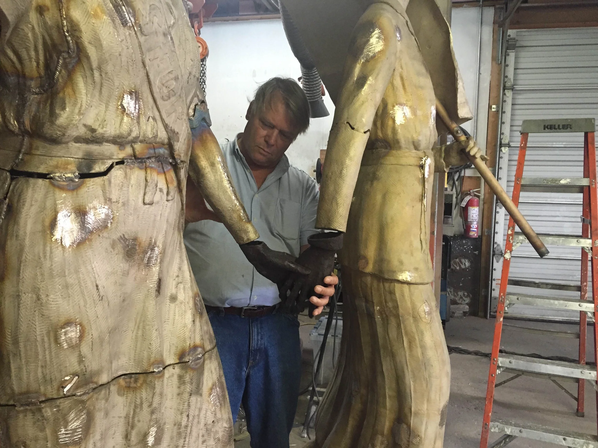 A man working on a gold-colored bronze sculpture in a workshop, surrounded by tools, scaffolding, and a fire extinguisher.