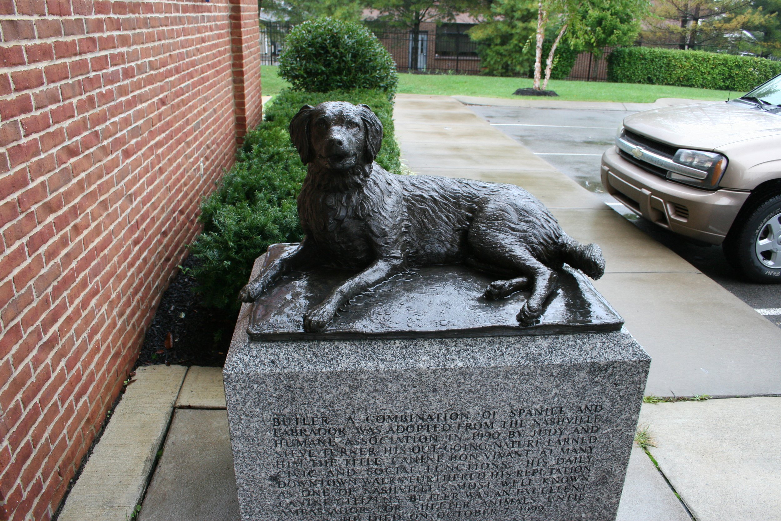 Bronze sculpture of a reclining dog on a stone pedestal, located outdoors next to a brick wall, with a parking lot and trees in the background.