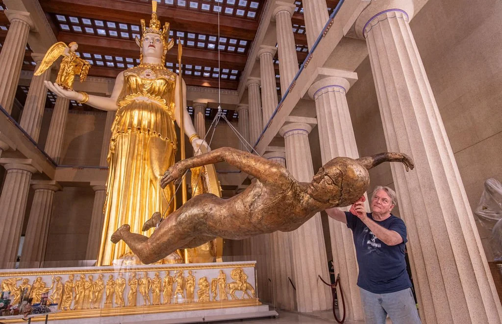 A man is standing next to a large, suspended sculpture of a human figure made of a bronze-like material, which is in front of an ornate, gold-colored statue of a goddess holding a bird, in a grand hall with tall columns and a wooden ceiling.