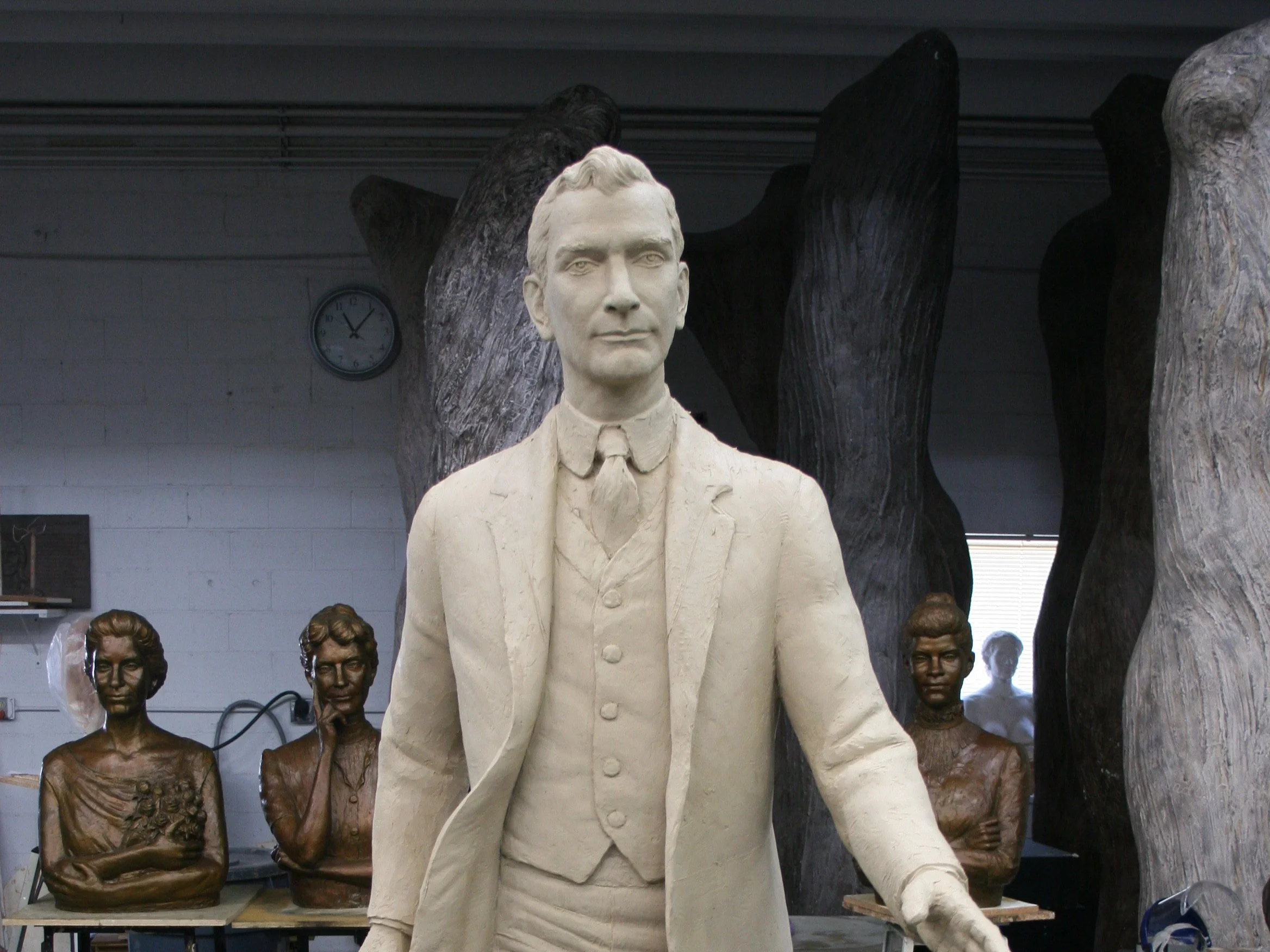 A white statue of a man in a suit and tie, standing with a confident expression. Several smaller bronze bust sculptures of women are arranged behind him on a shelf in a workshop or studio setting.