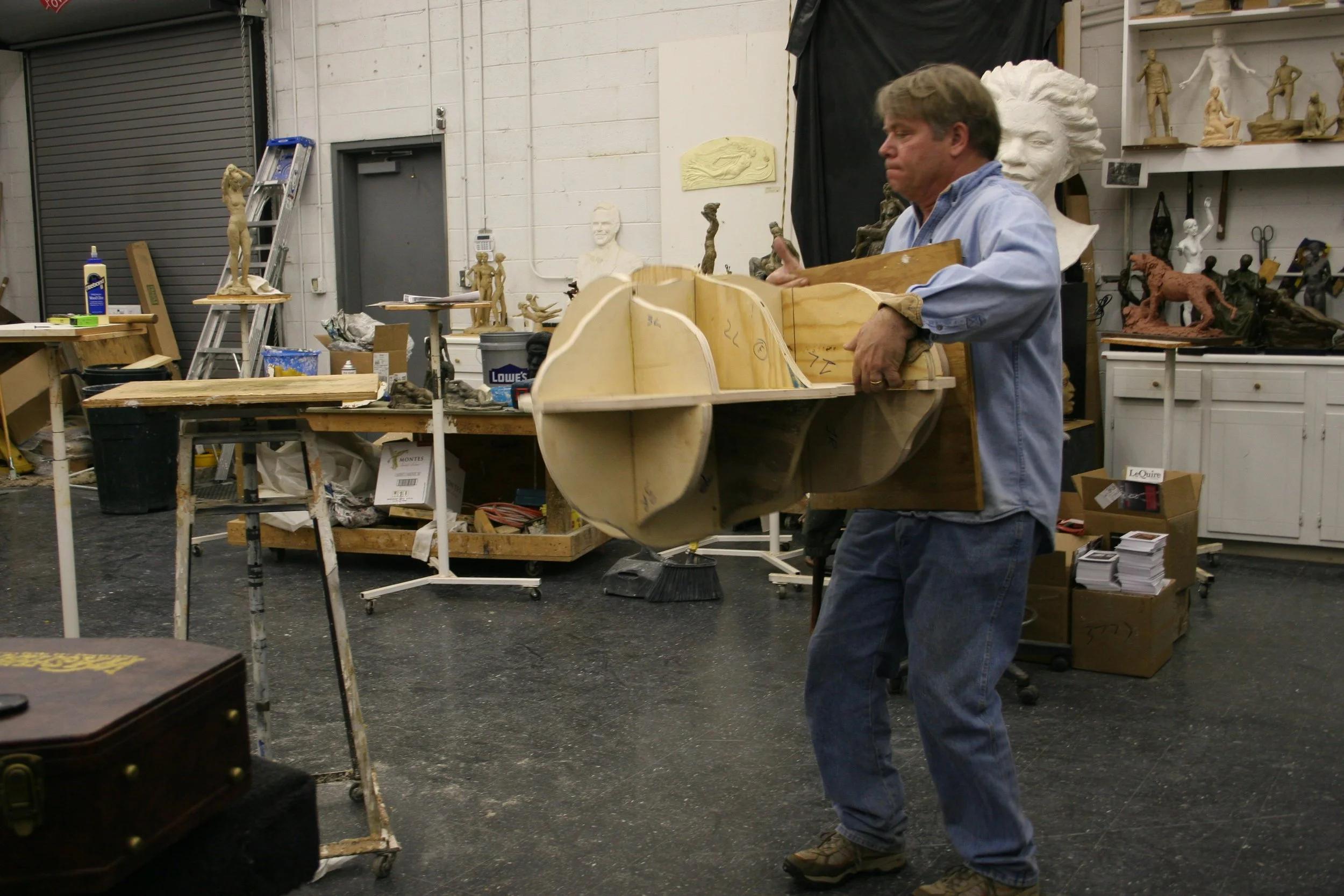 A man in a blue shirt working on a large wooden sculpture in a workshop filled with art sculptures, tools, and materials.
