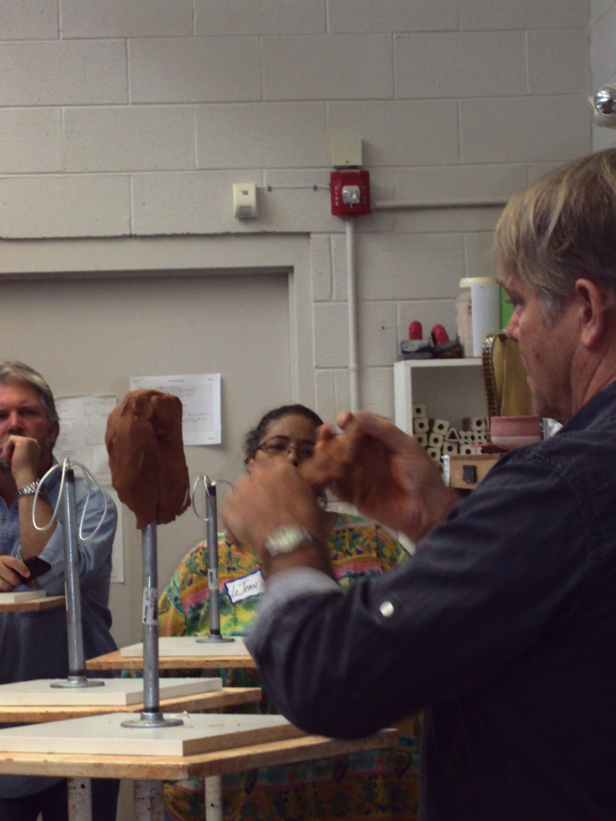 Group of people participating in a professional or educational workshop, sitting at tables with materials, and engaging in discussion, in a room with beige walls and shelves.