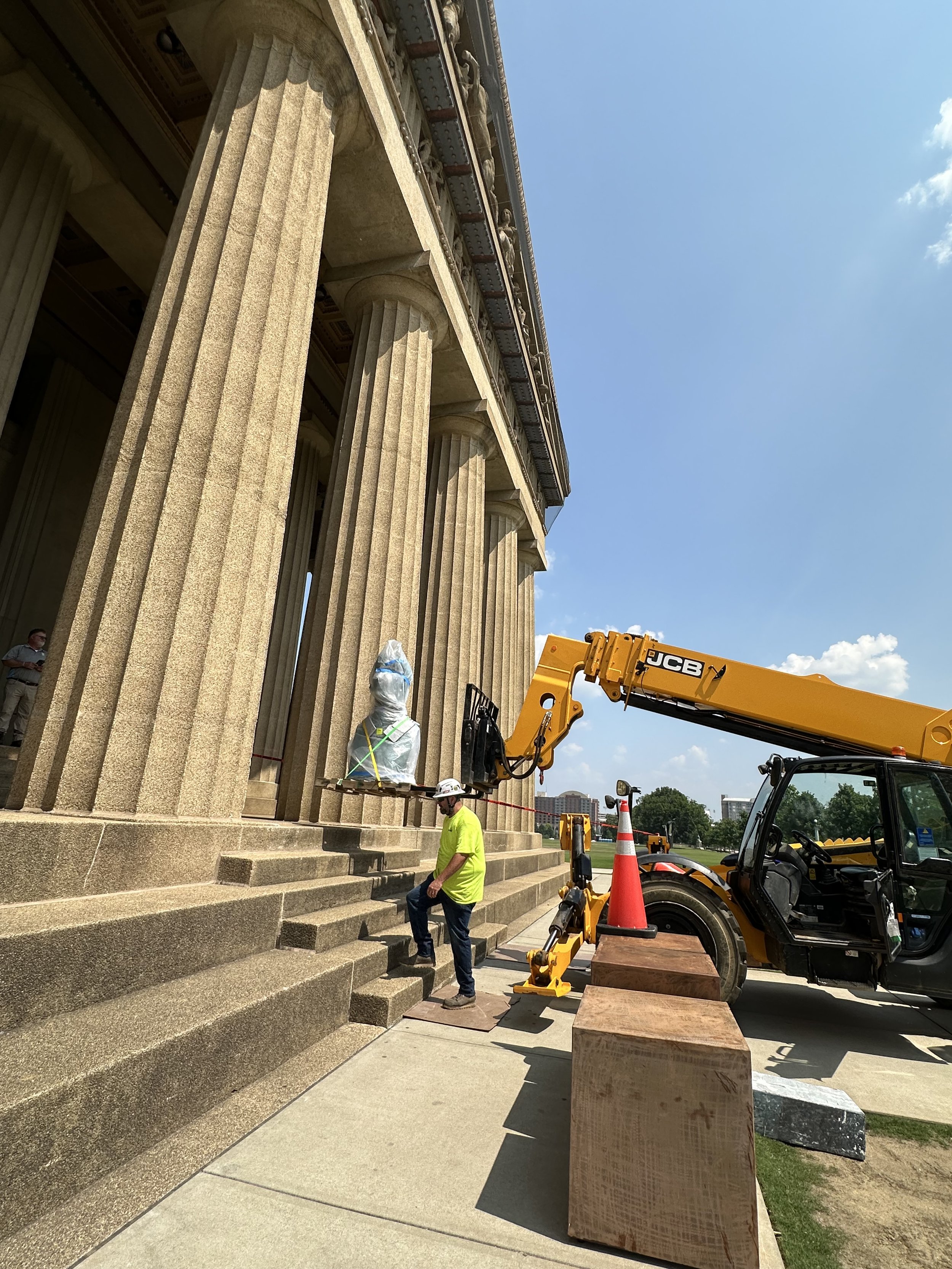 Workers in construction gear working on the steps of a large, classical stone building with tall columns under a clear, blue sky. Construction equipment, including a JCB crane, is present.