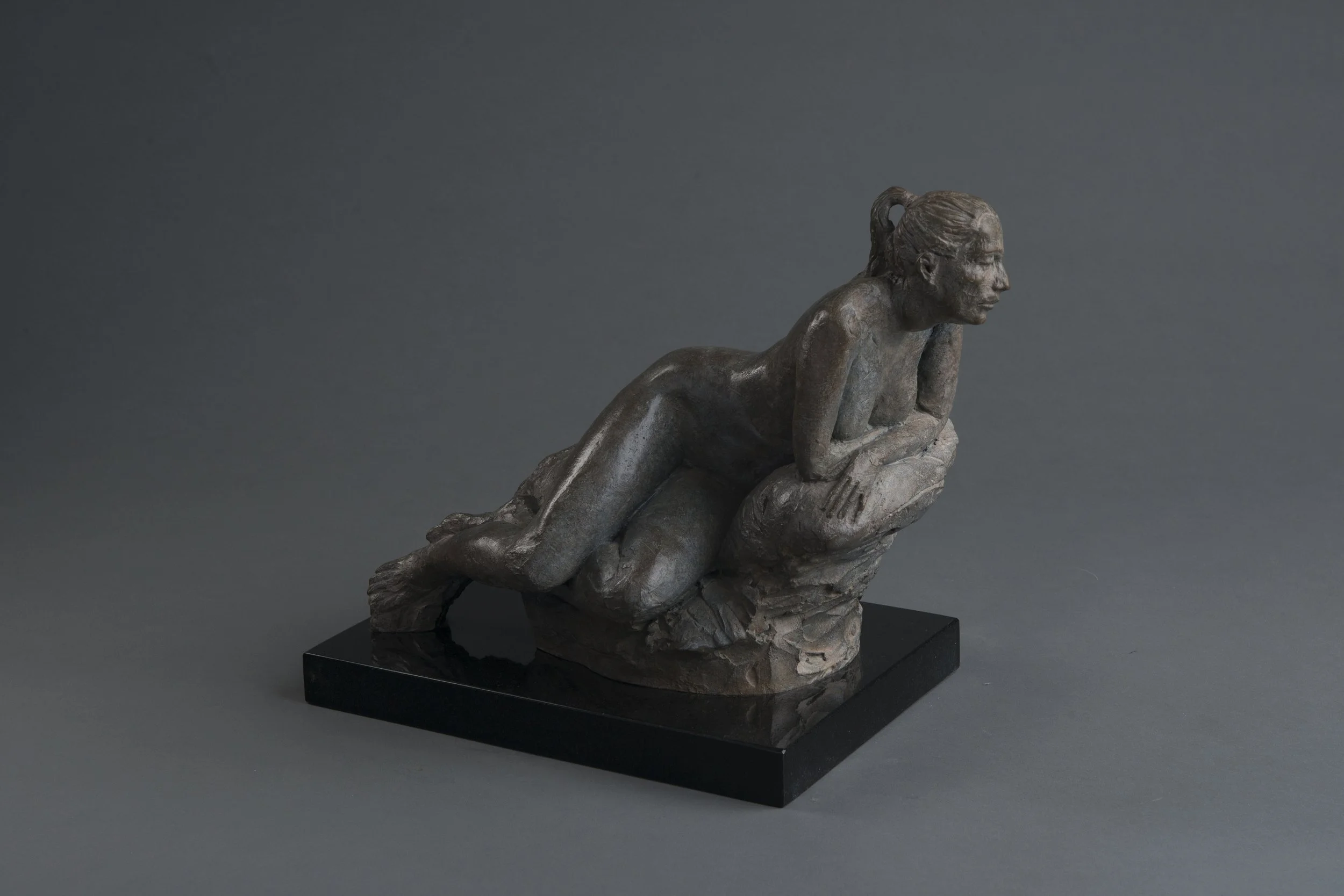 Bronze sculpture of a woman crouching on a rock, resting her chin on her hand, set against a dark gray background.
