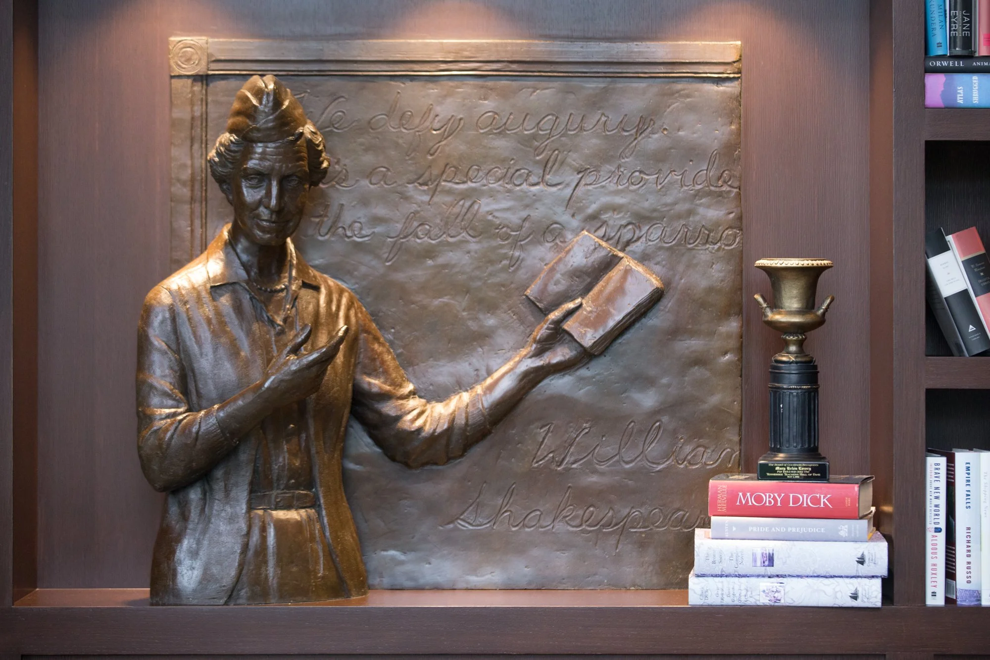 Bronze sculpture of a woman with her hair in a bun, wearing a blazer and pearl necklace, holding a book in her left hand and gesturing with her right hand. Behind her is a plaque with cursive writing. To the right, there are books on a shelf, including one titled "Moby Dick" and a trophy with a black base and gold top.