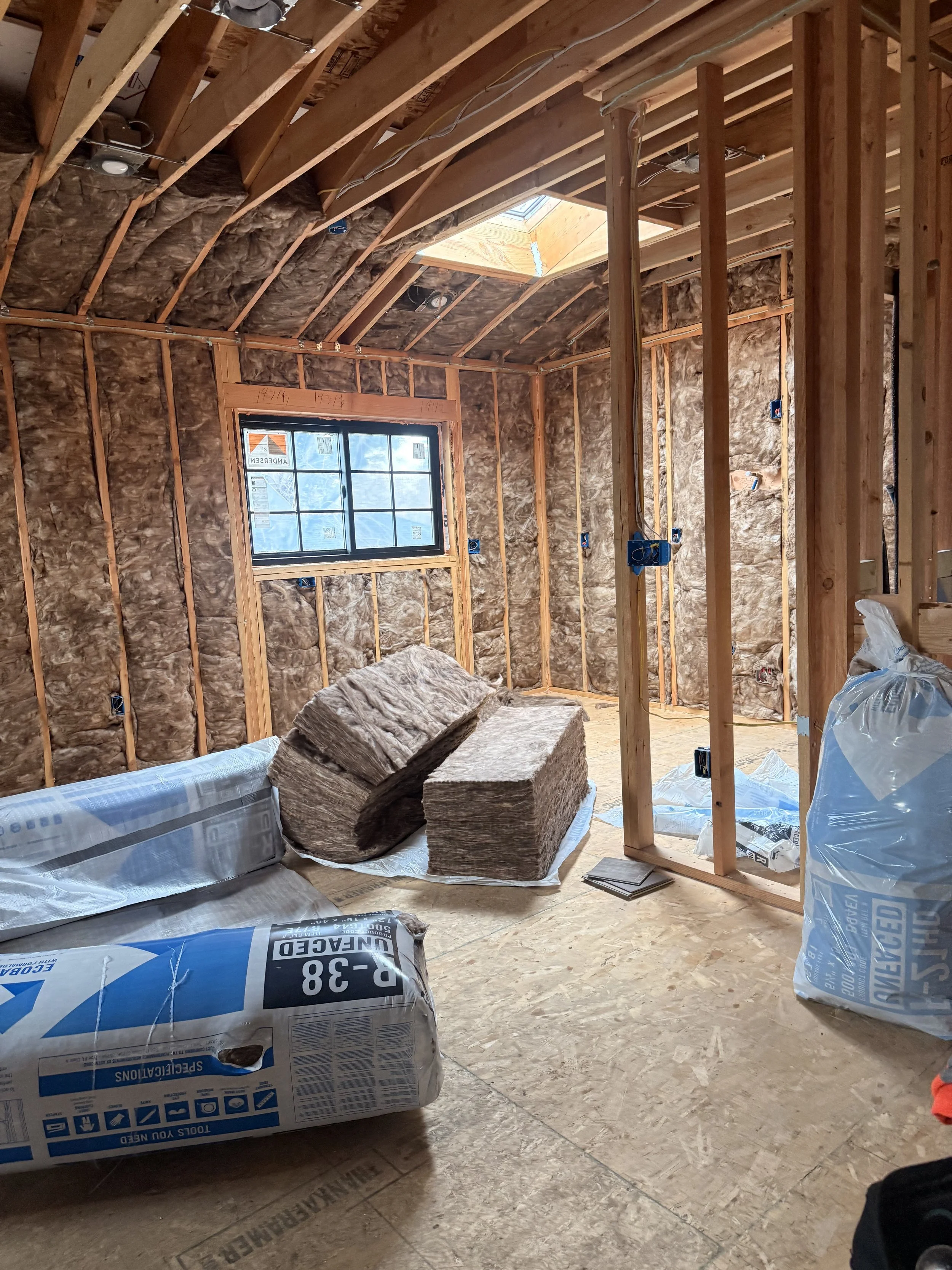 Interior of a room under construction with exposed wooden framing, insulation, a window, and construction materials on the floor.
