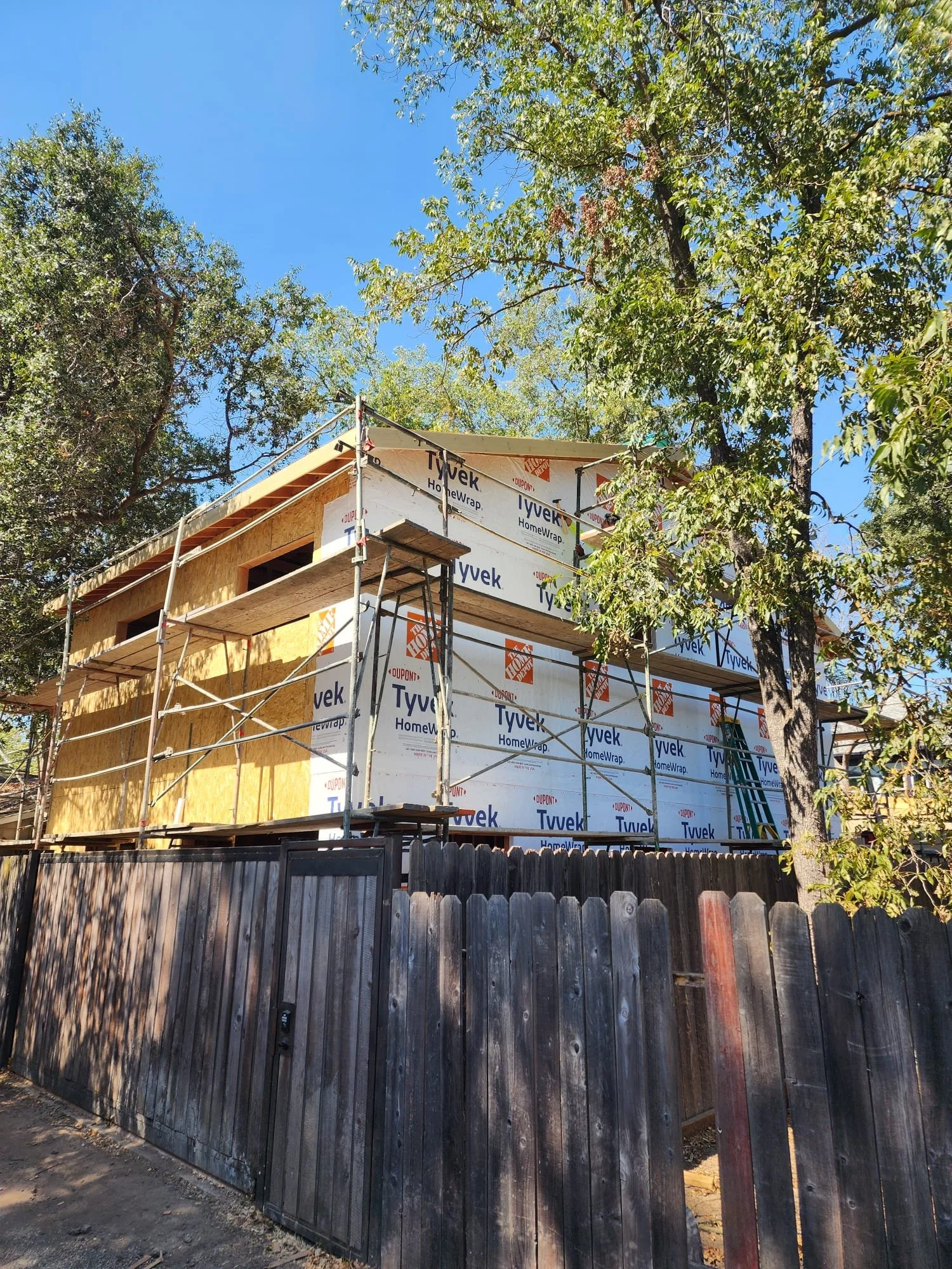 Under construction house with Tyvek HomeWrap on exterior, wooden framing visible, surrounded by a wooden fence, trees in background, clear blue sky.