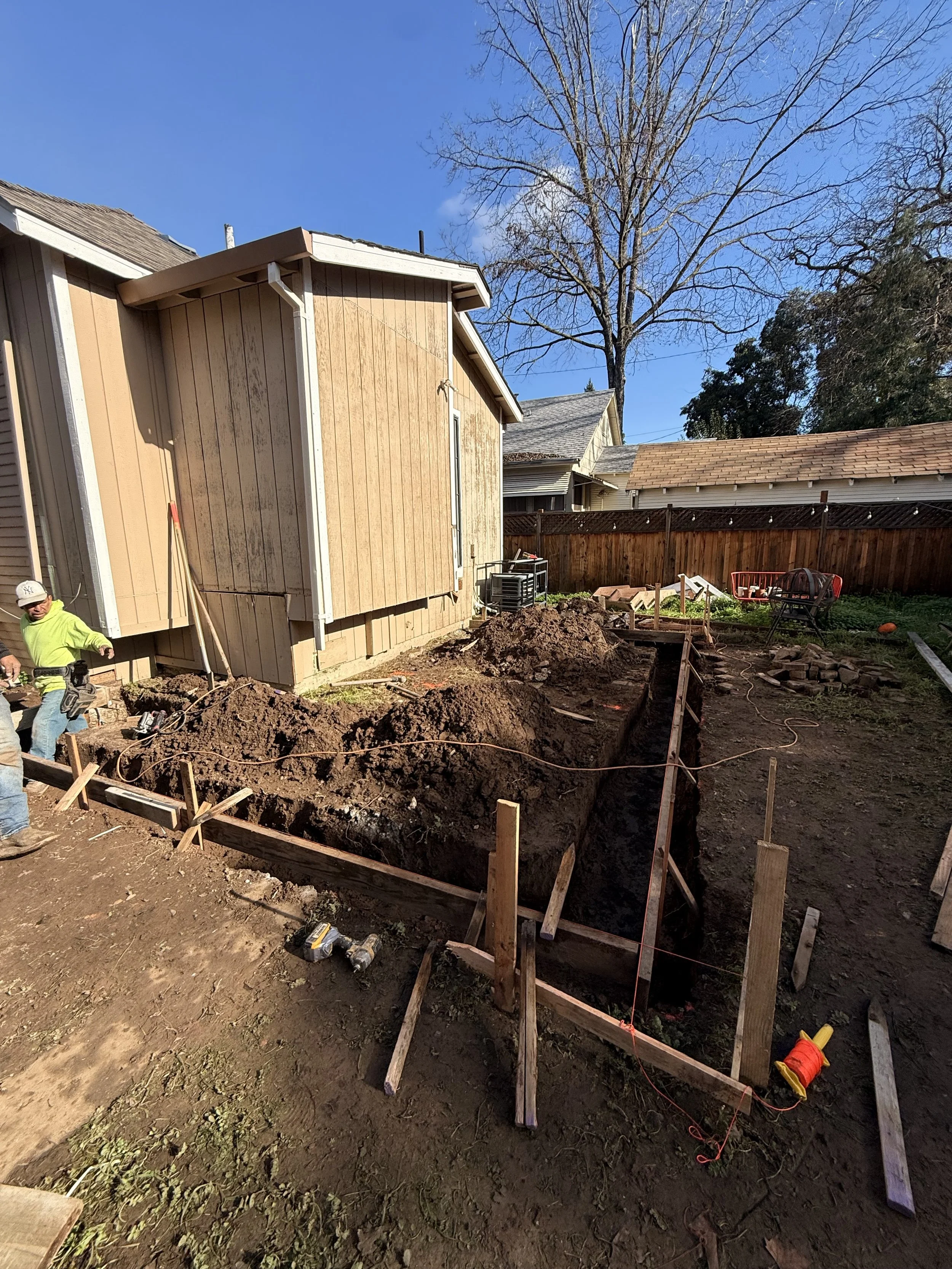 Construction site in a backyard with a trench being dug, wooden stakes and framing, and two workers wearing safety gear; house with vertical wooden siding in the background, leafless tree, and clear blue sky.