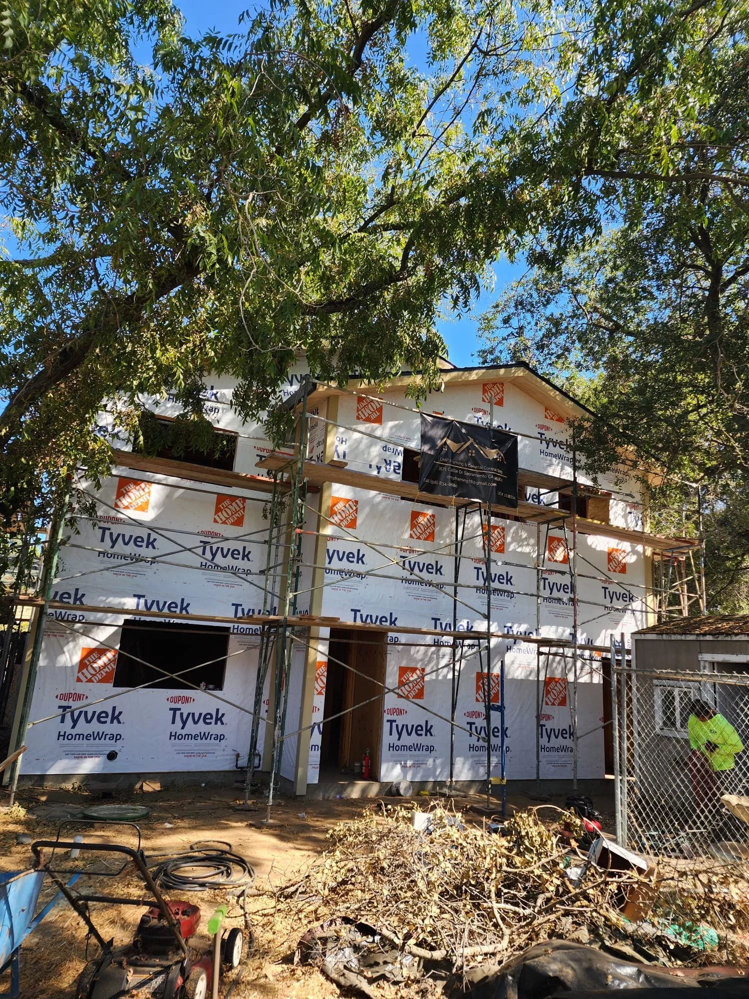 Under construction house wrapped in Tyvek HomeWrap material with scaffolding and construction workers present, surrounded by trees and construction tools.