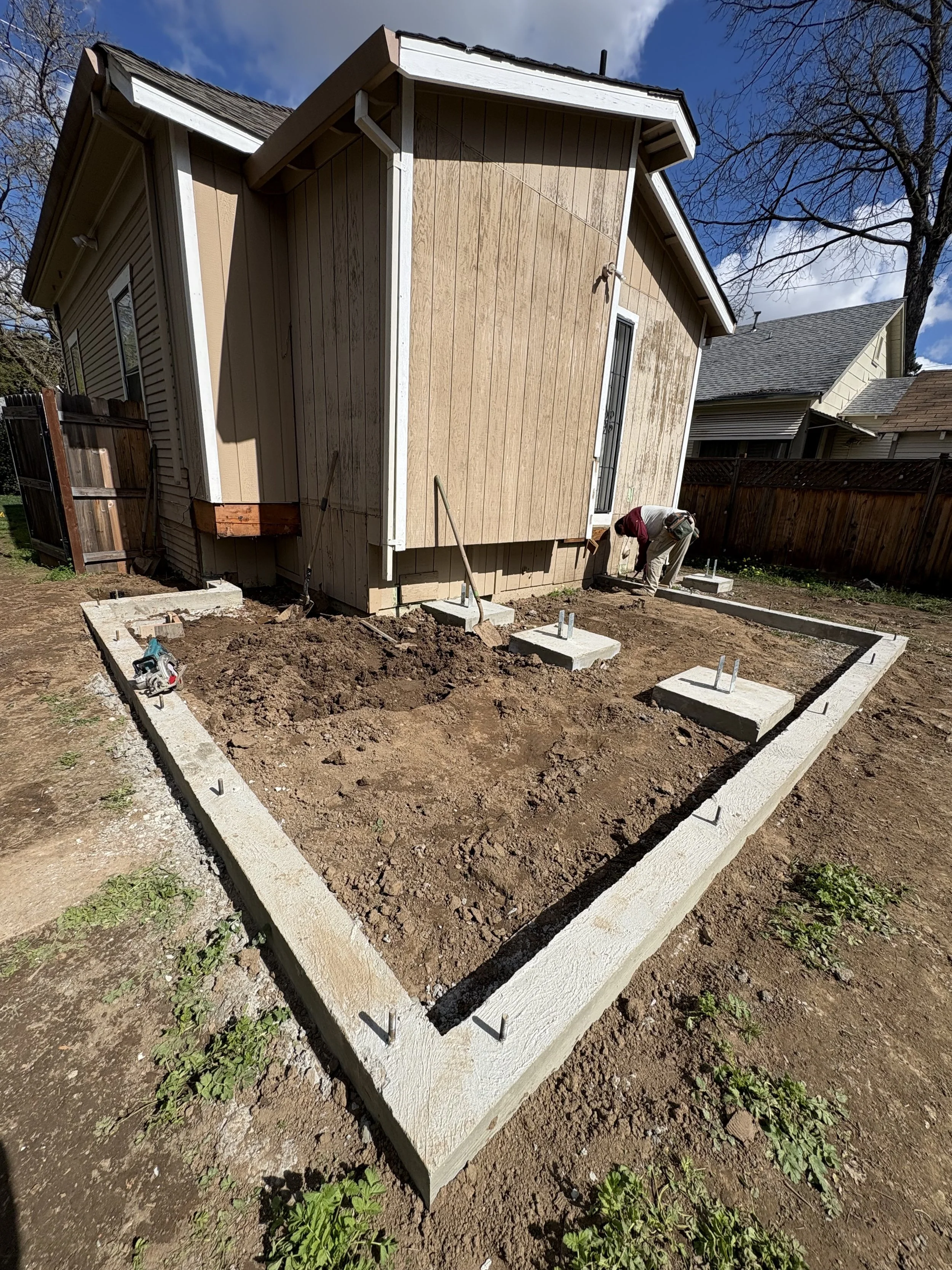 Construction site with a house, workers, and foundation being prepared with concrete blocks and rebar for a new structure in a backyard, under a blue sky with some clouds.