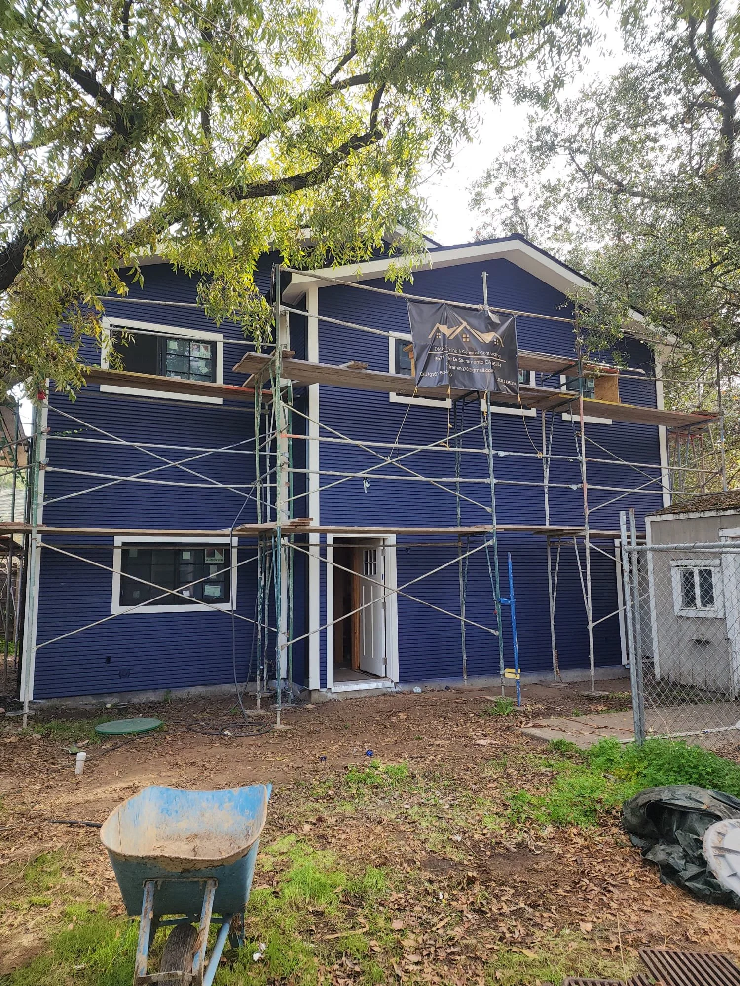 A two-story house under construction with blue exterior siding, scaffolding around it, and a wheelbarrow in the yard.