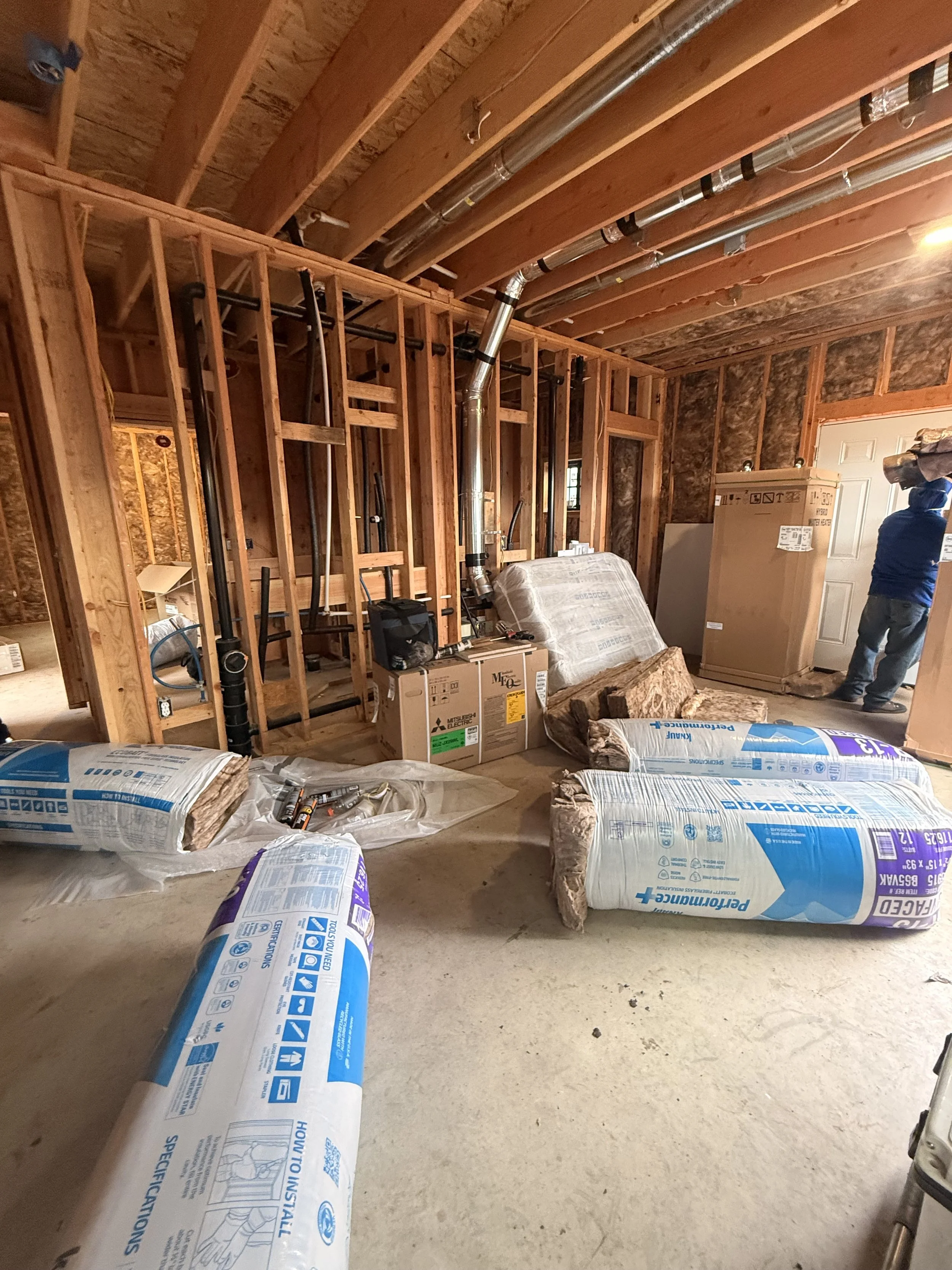 Interior of an unfinished room with exposed wooden framing, pipes, insulation materials, and construction supplies, with a person standing near a door in the background.