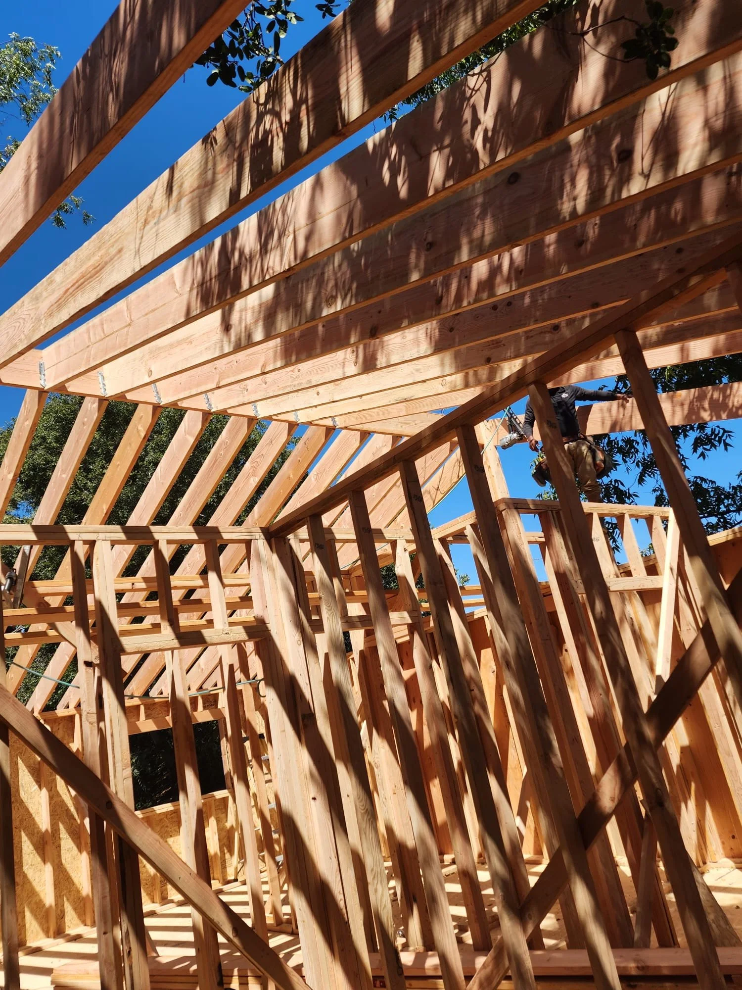 View of a wooden house frame under construction with a worker standing on a platform, surrounded by trees and blue sky.