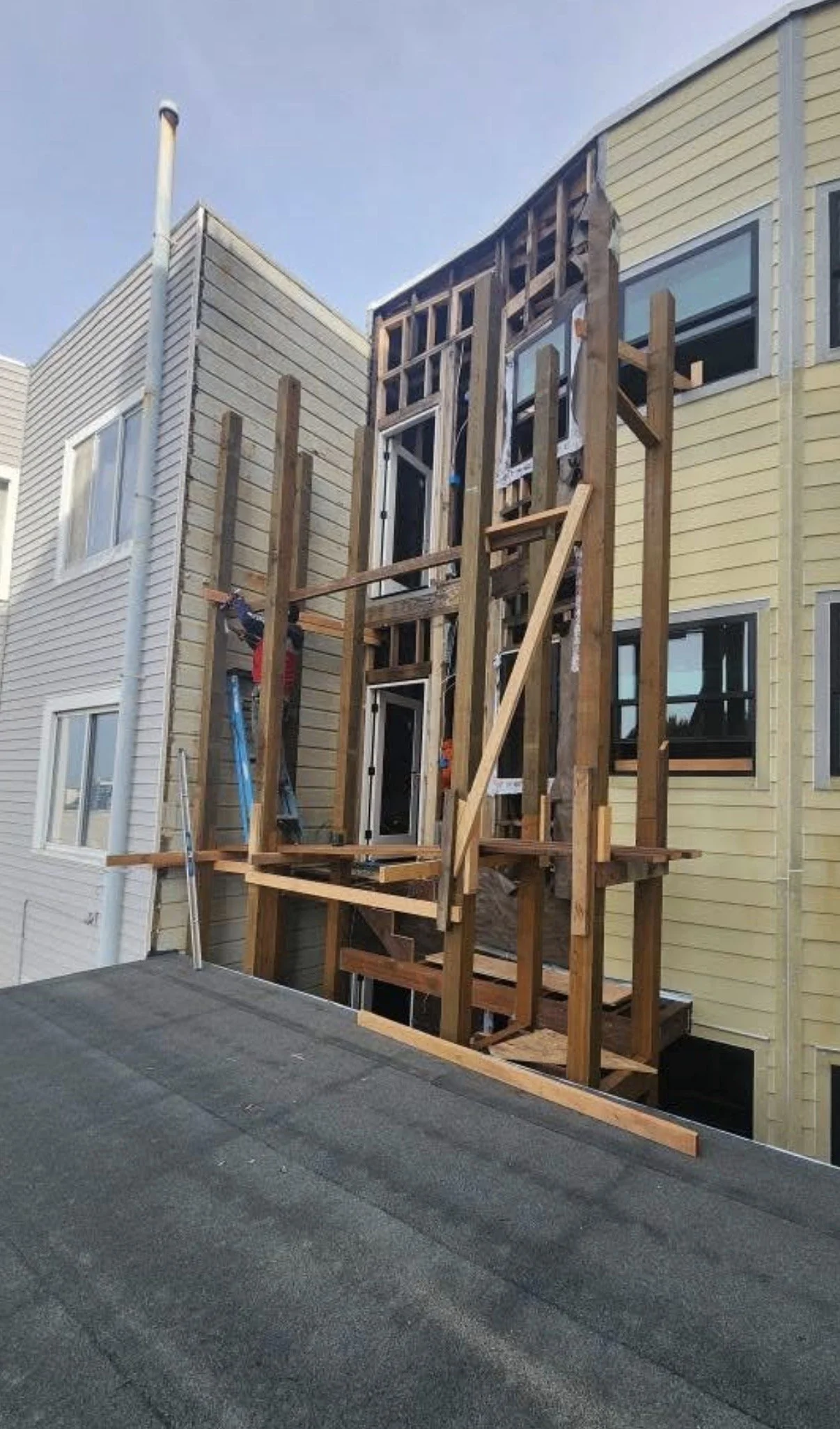 Construction site with a wooden framework for an extension or balcony on a multi-story building, surrounded by neighboring buildings with siding.