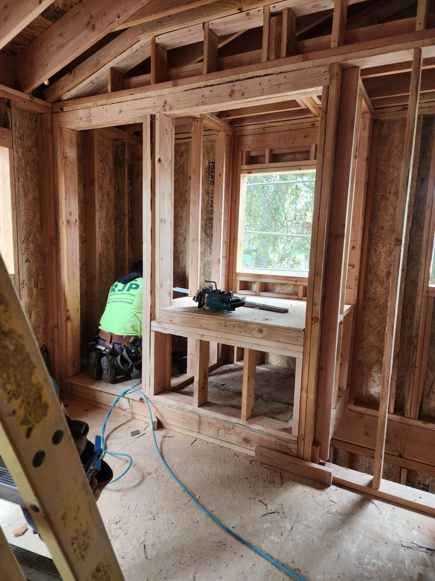 Interior of a house under construction with exposed wooden framing, a window with a view of trees outside, a work table with a power saw, and a worker installing or inspecting the structure.