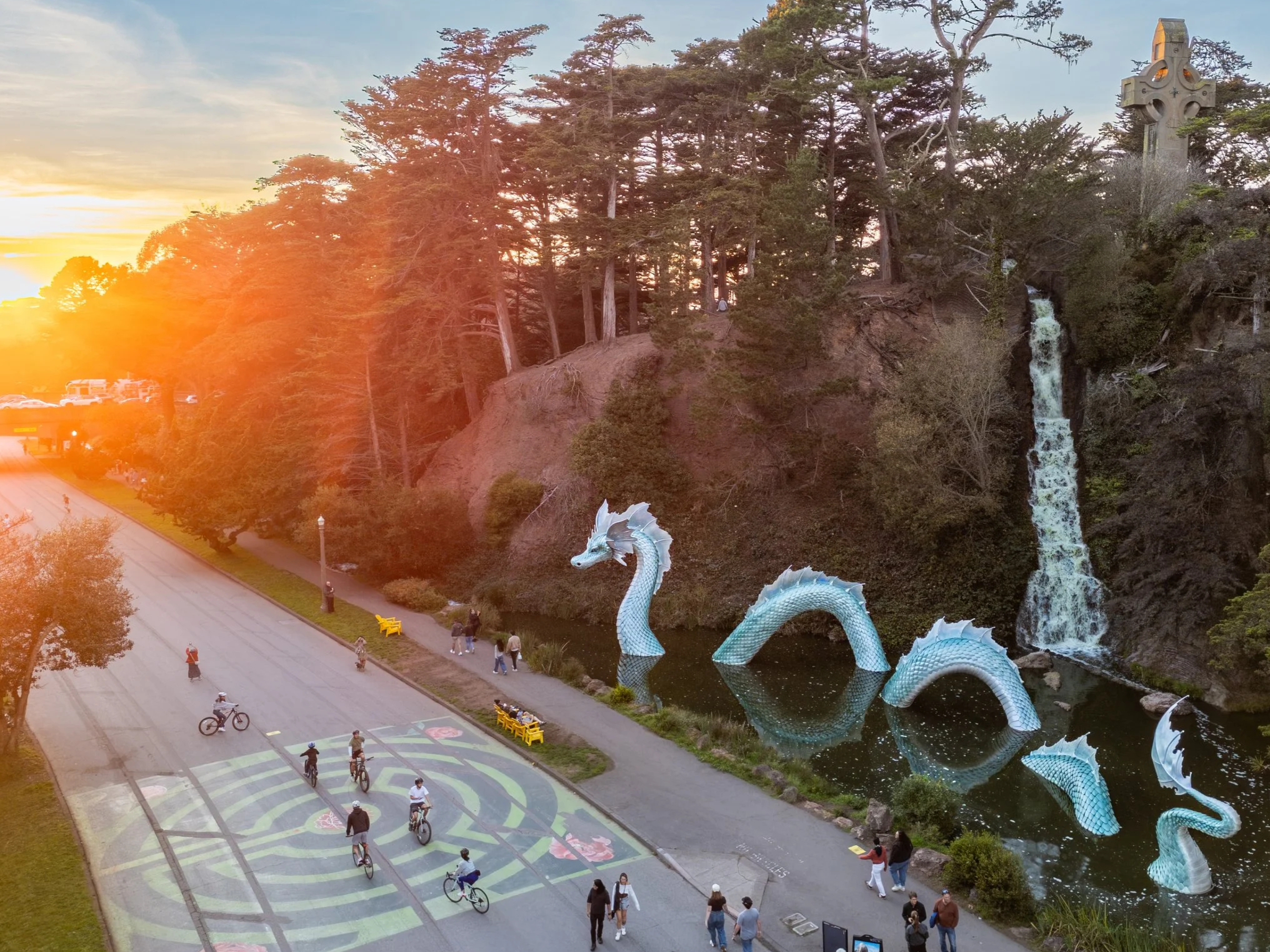 Sunset over a park with a large sculptures of a sea serpent and waterfall, people cycling and walking, and a cloudy sky.