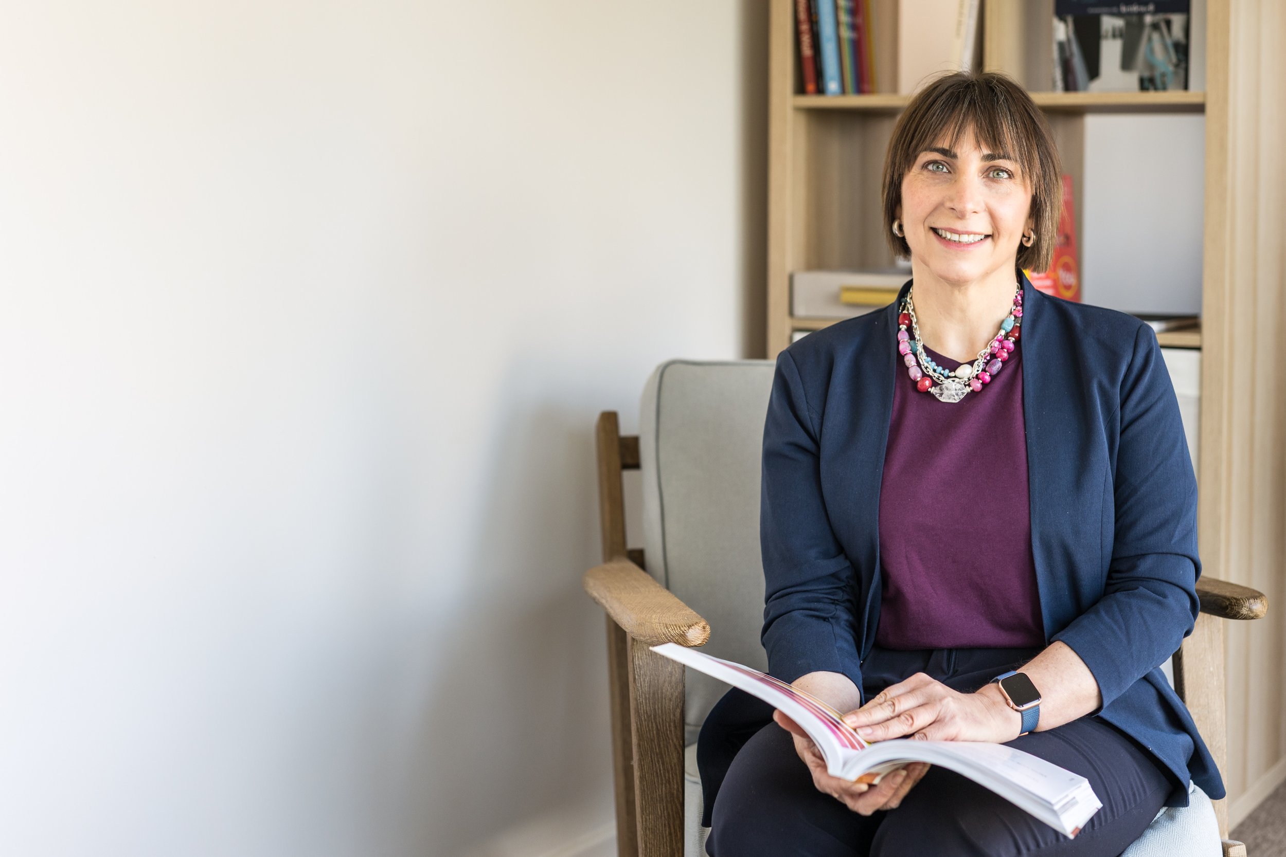 A woman sitting on a wooden chair, smiling, holding a magazine or booklet, in a room with bookshelves in the background.