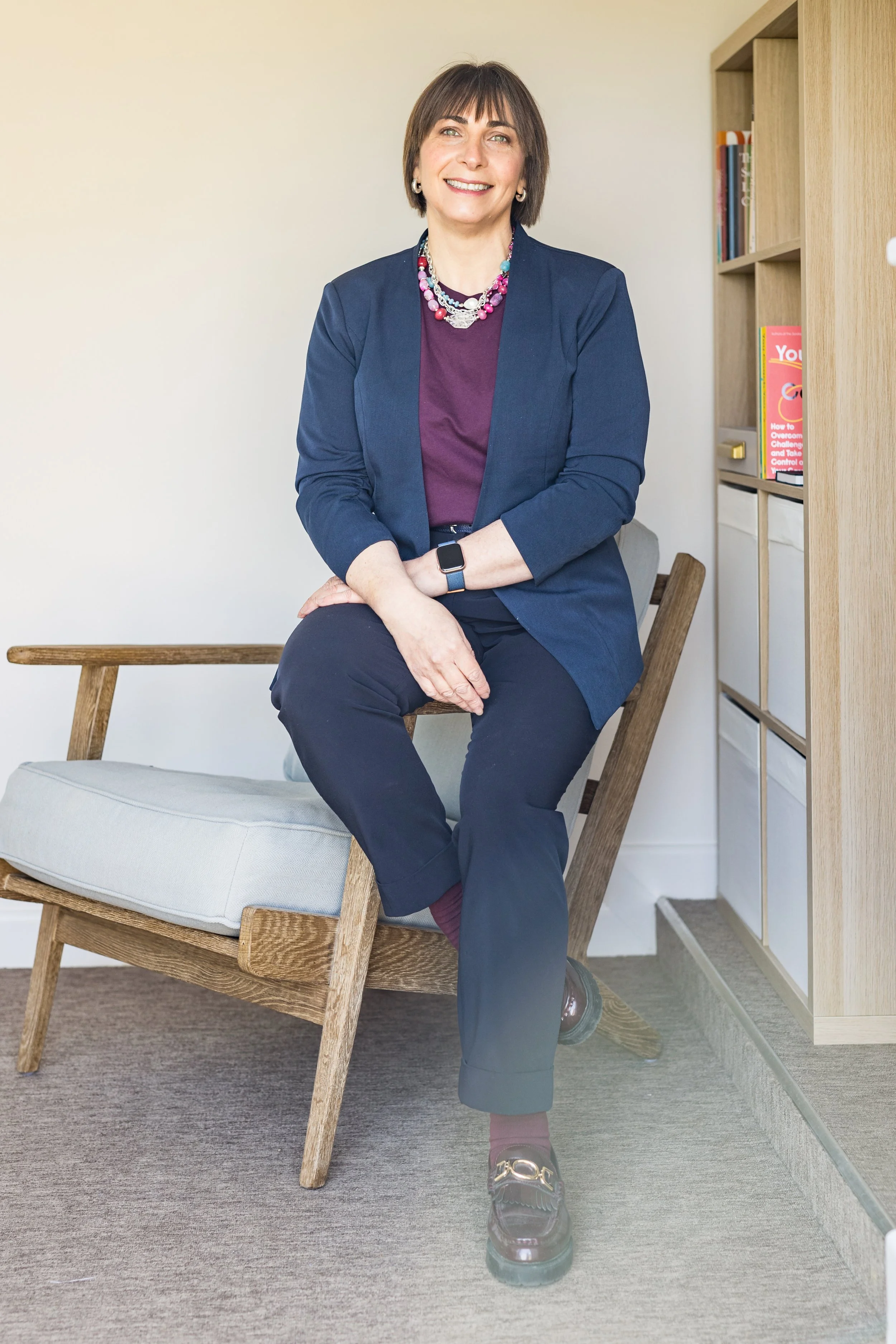 A woman with short dark hair is sitting on a wooden chair with a gray cushioned seat. She is wearing a navy blazer, maroon pants, black shoes, and a colorful beaded necklace. She has a smartwatch on her left wrist and is smiling at the camera. Behind her is a bookshelf with books and storage bins, and the room has light-colored walls and gray carpet.