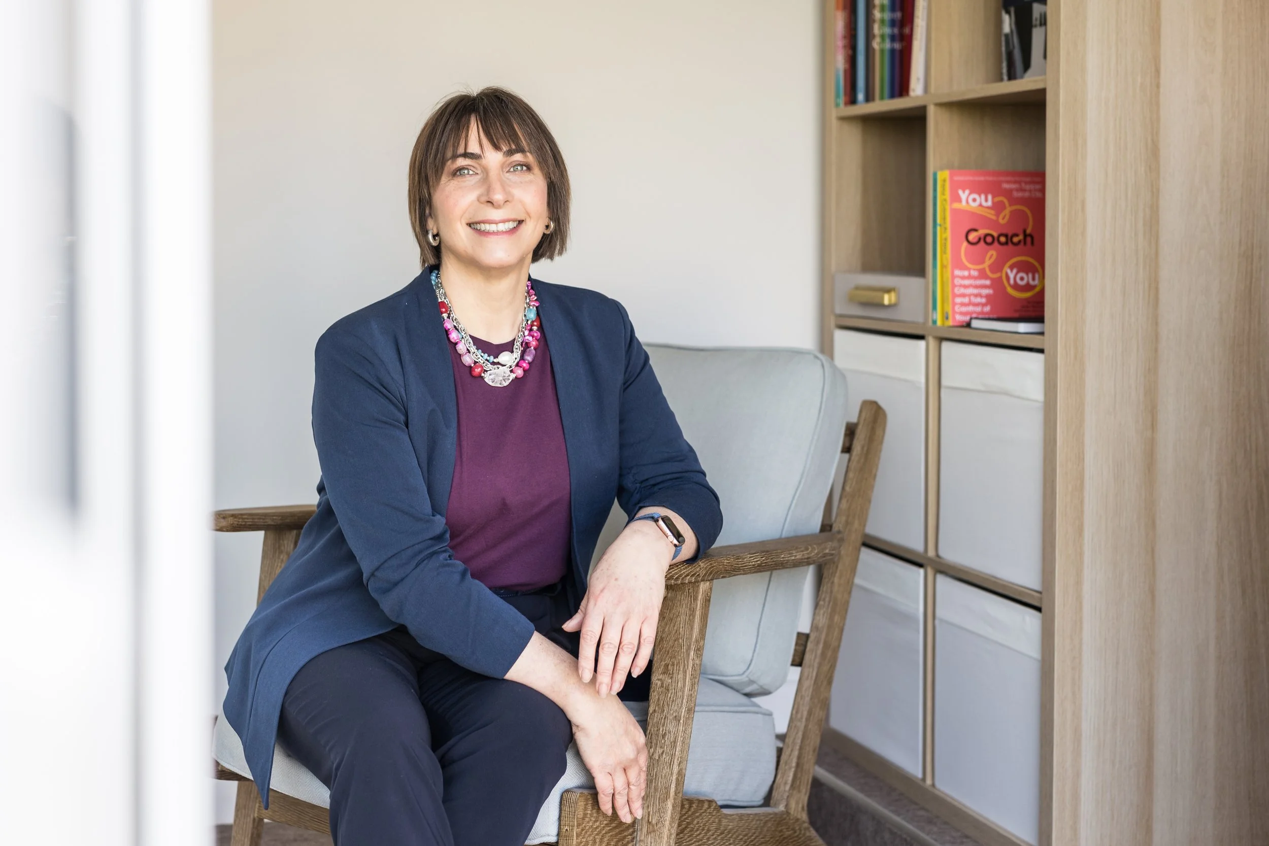 A woman with short brown hair and blue eyes sitting on a wooden chair with a grey cushion, smiling, in a room with a light-colored wall and a bookshelf filled with books behind her.
