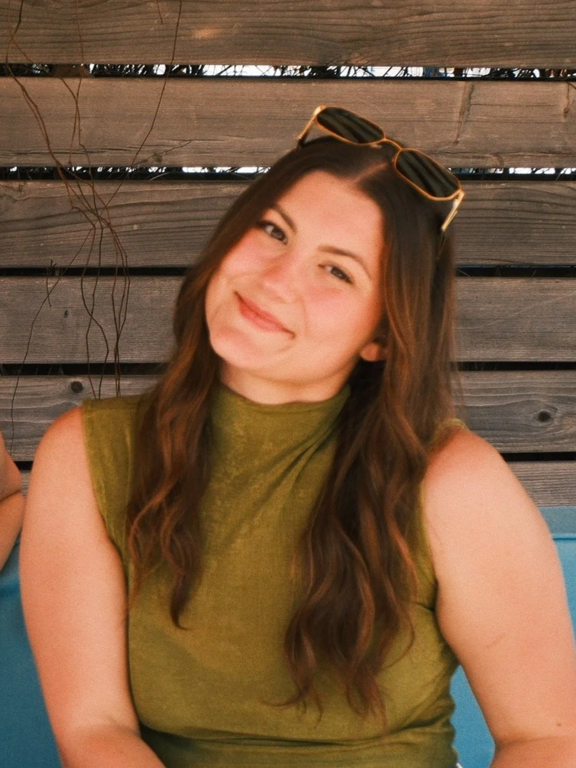 A young woman with long, wavy brown hair wearing sunglasses on her head and a sleeveless olive green top, sitting against a wooden fence with some vines.