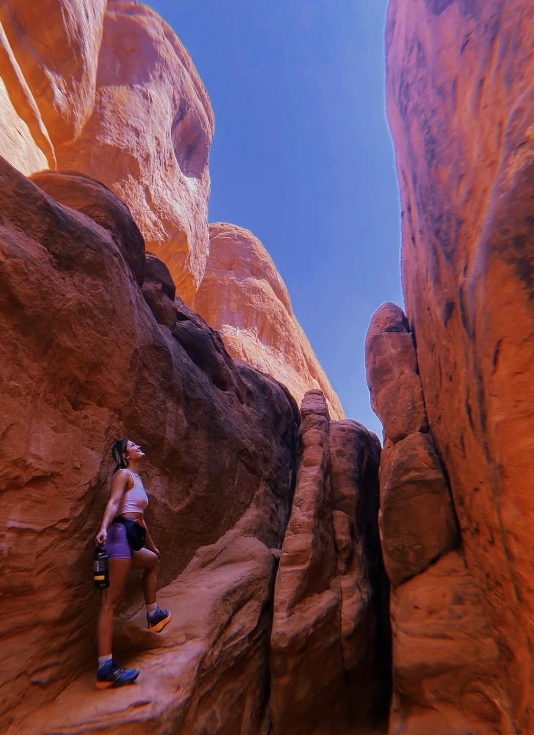 A woman standing on a narrow rock ledge in a deep, red sandstone canyon looking up at the tall canyon walls and clear blue sky.