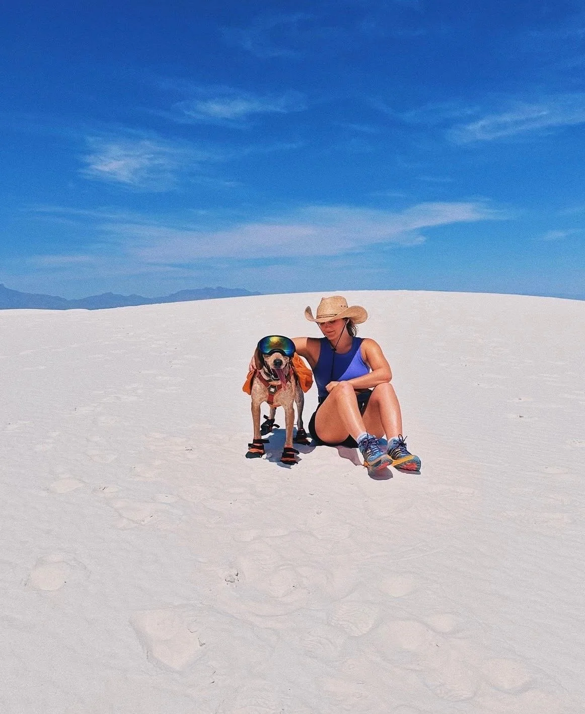 Woman and dog sitting on white sandy terrain under blue sky, woman wearing large sunhat and athletic clothing, dog wearing sunglasses and shoes, both enjoying sunny day outdoors.