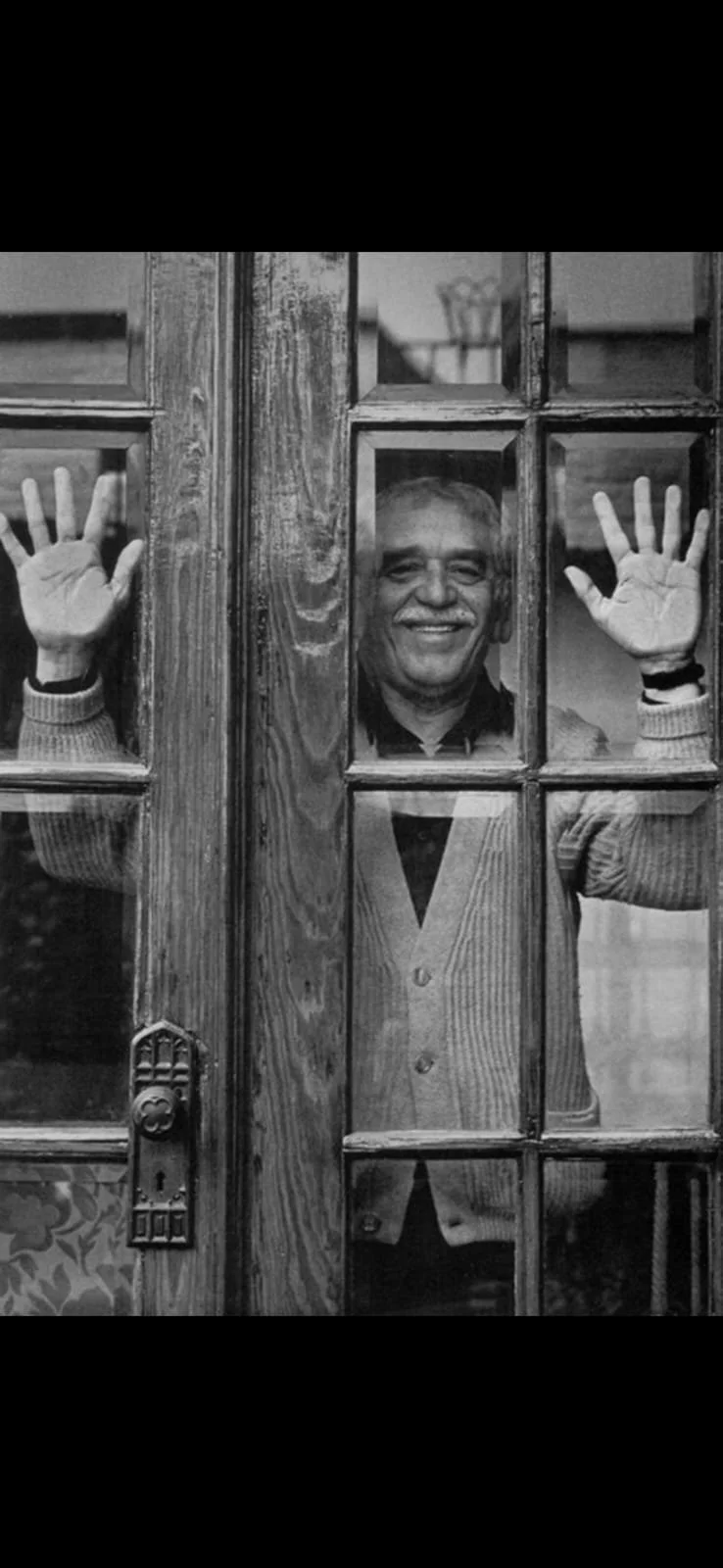 Hombre sonriendo detrás de una puerta de cristal, saludando con las manos, vista en blanco y negro.