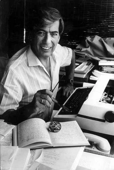 Un hombre con cabello oscuro y sonrisa, sentado en un escritorio con una computadora, libros y objetos personales, sosteniendo un lápiz en la mano.