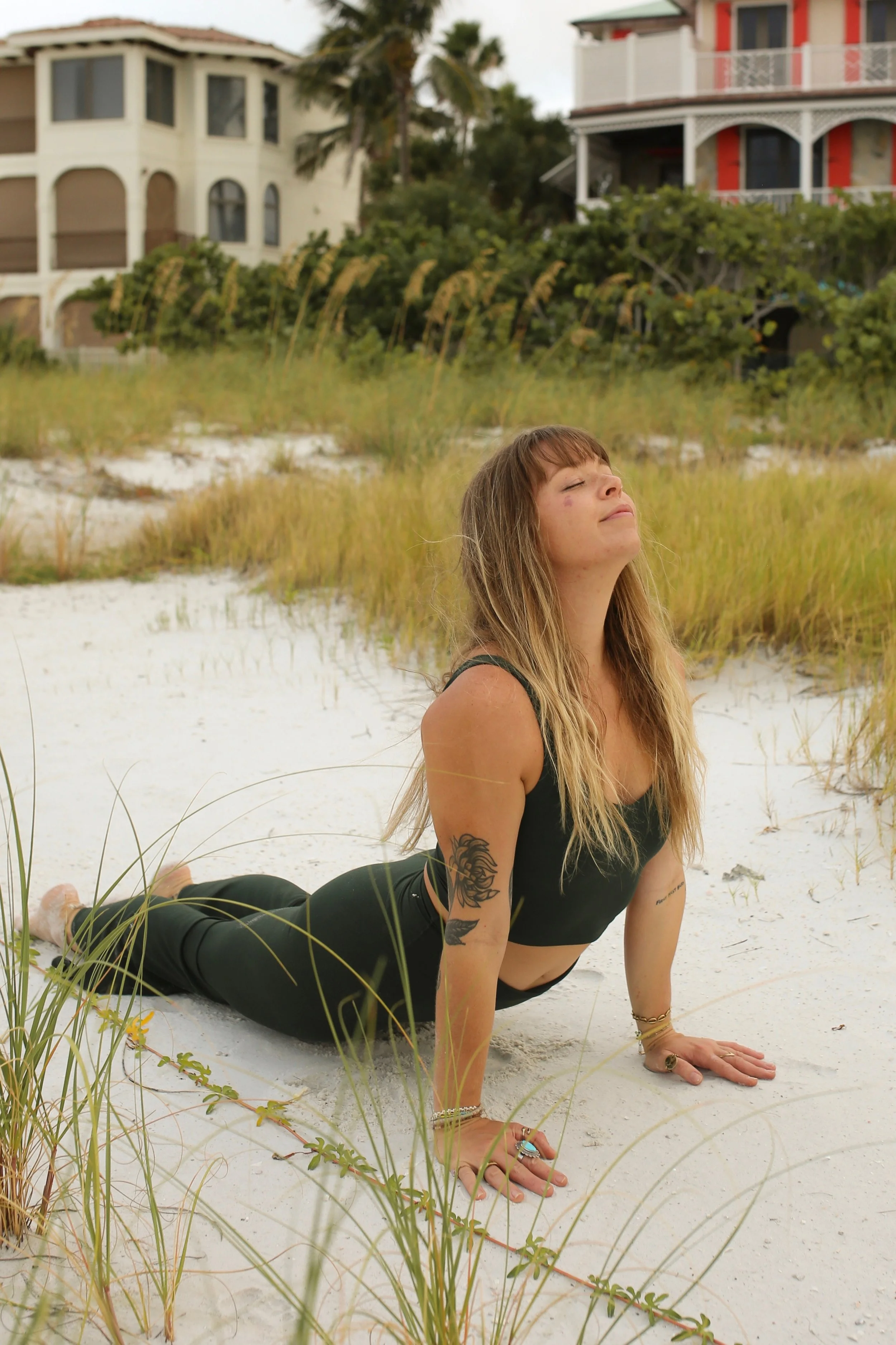 A woman practicing yoga on the beach, in a cobra pose, with houses and palm trees in the background.