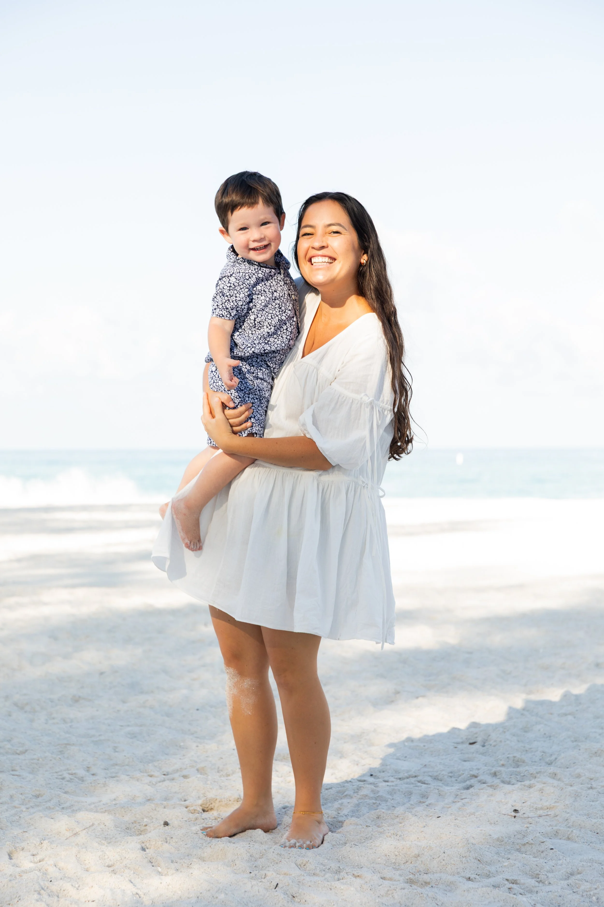A woman with long dark hair, wearing a white dress, is holding a smiling young boy with dark hair, dressed in a patterned shirt, at the beach with sand and the ocean in the background.