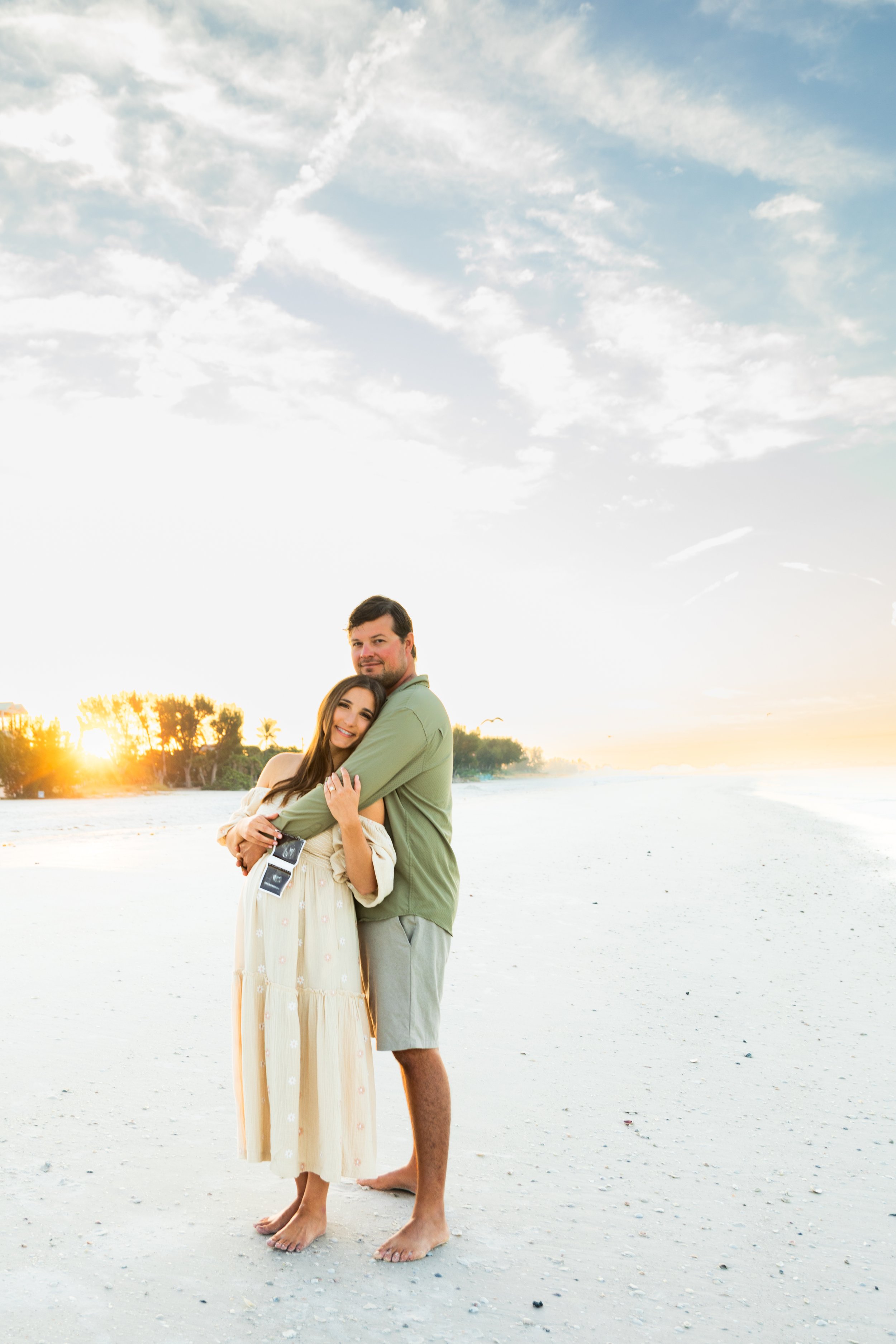A smiling man and woman embrace on a beach during sunset, with the sky partly cloudy and trees in the background.