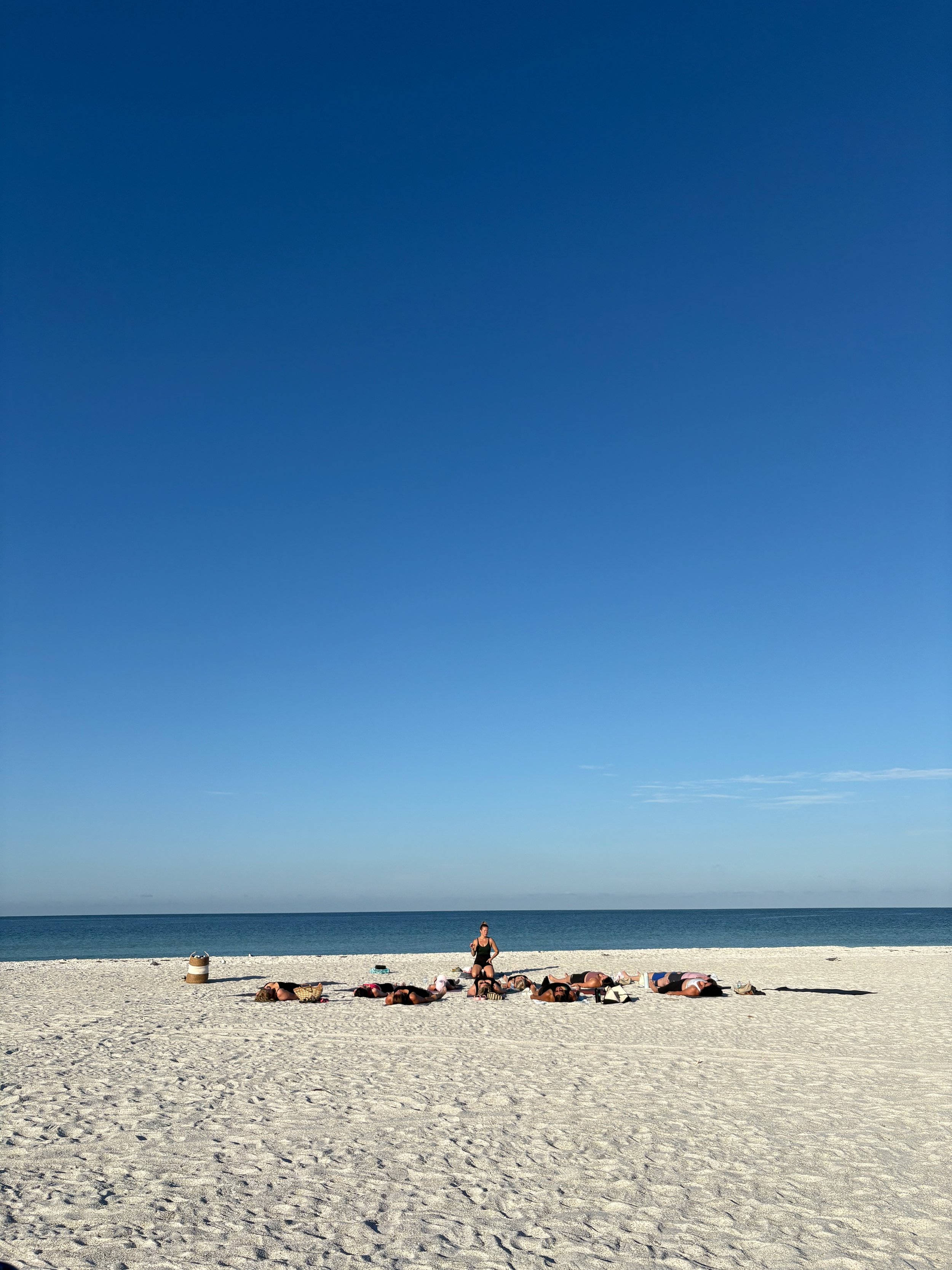 A woman practicing yoga on a sandy beach with a group of people lying down in a circle around her. The ocean and blue sky with some clouds are in the background.