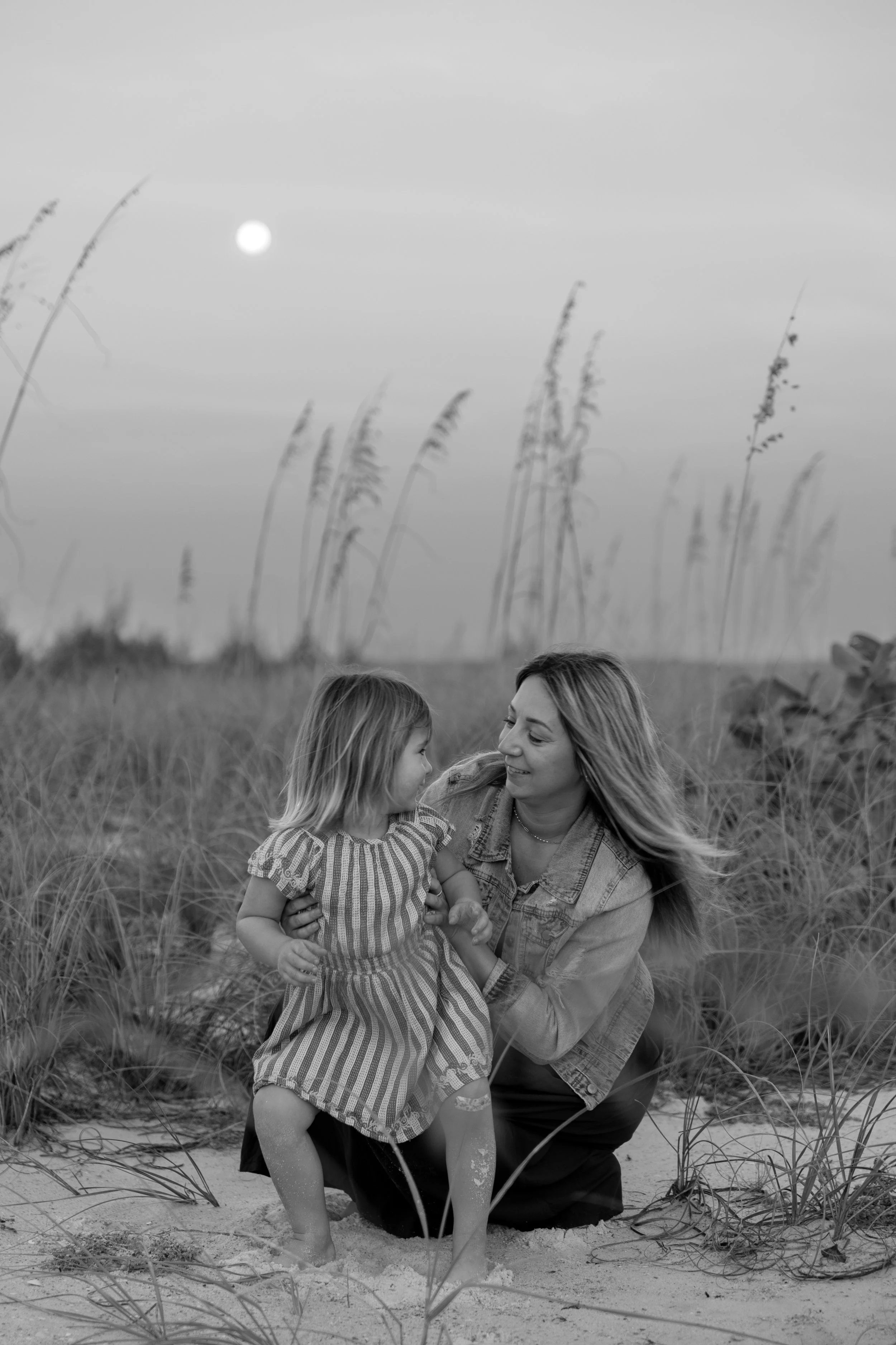 A woman and a young girl are at the beach surrounded by tall grass. The woman is crouched down and smiling at the girl, who is looking at her. The scene takes place at sunset or sunrise, with a bright sun in the sky. The image is in black and white.