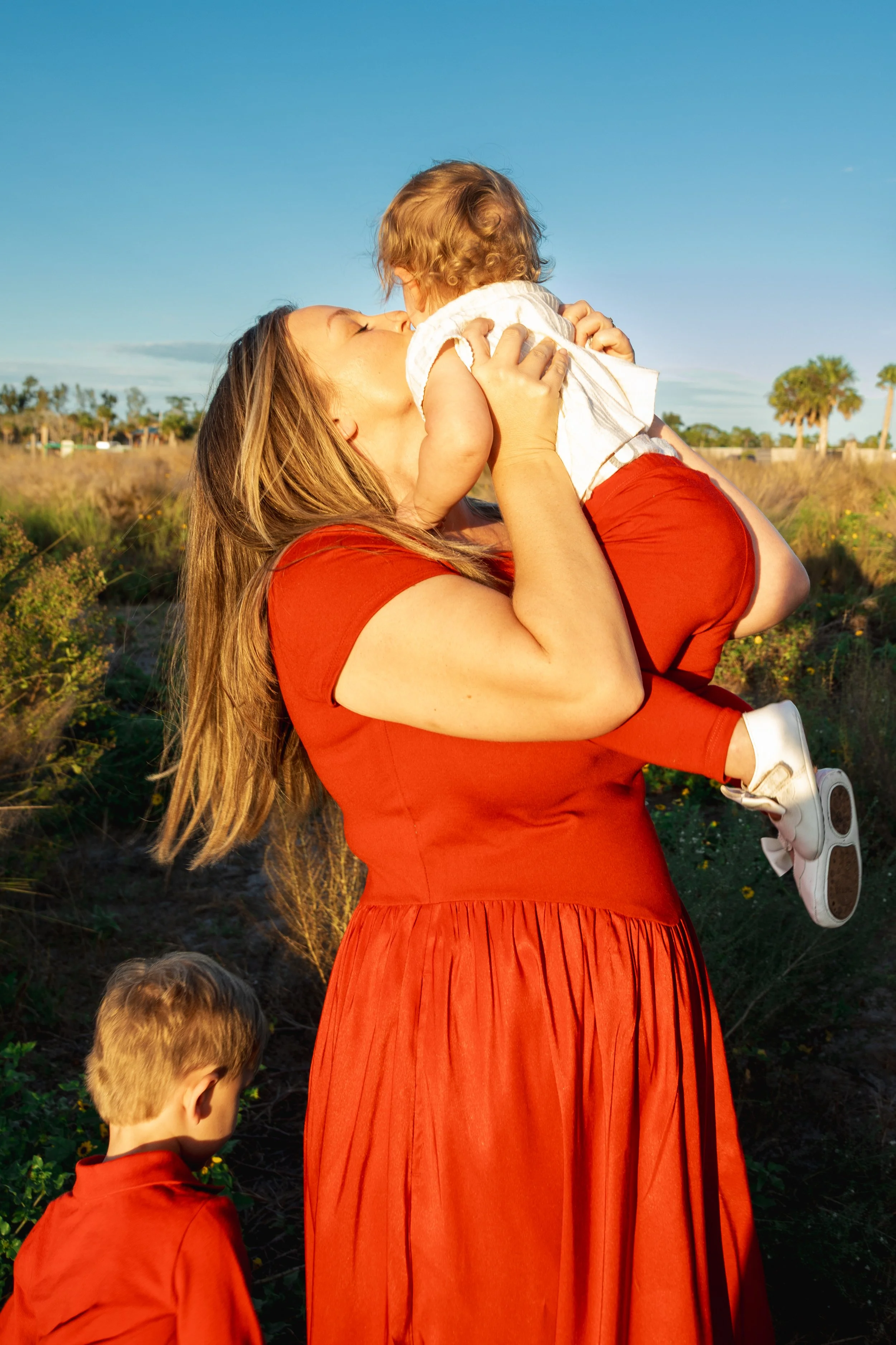 A woman with long hair in a red dress is holding a young child with curly hair, who is kissing her on the nose, outdoors on a sunny day with a clear blue sky and palm trees in the background.