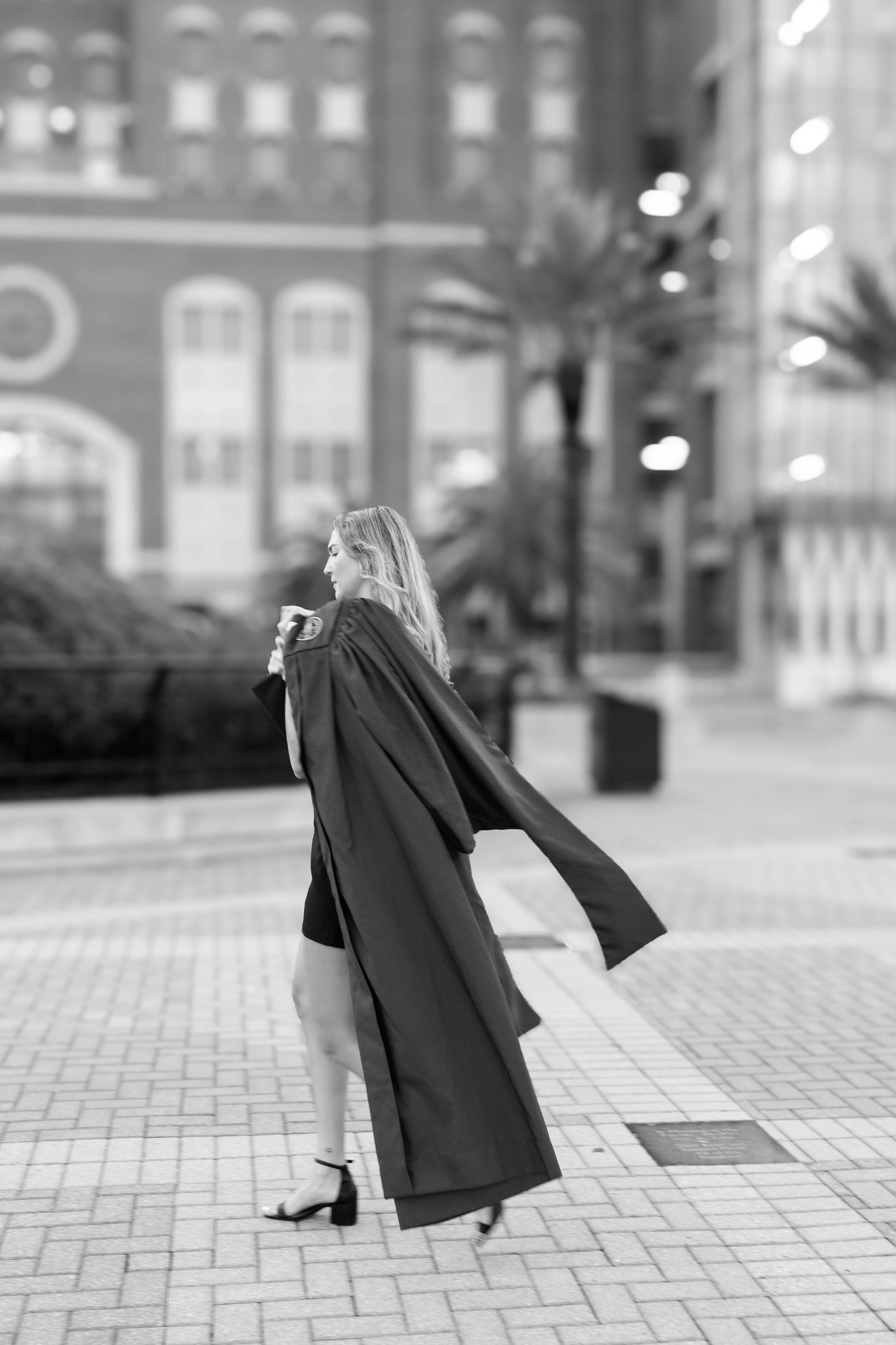 A woman walking outdoors in an urban setting, dressed in a dress and high heels, with a coat draped over her shoulders, captured in black and white.