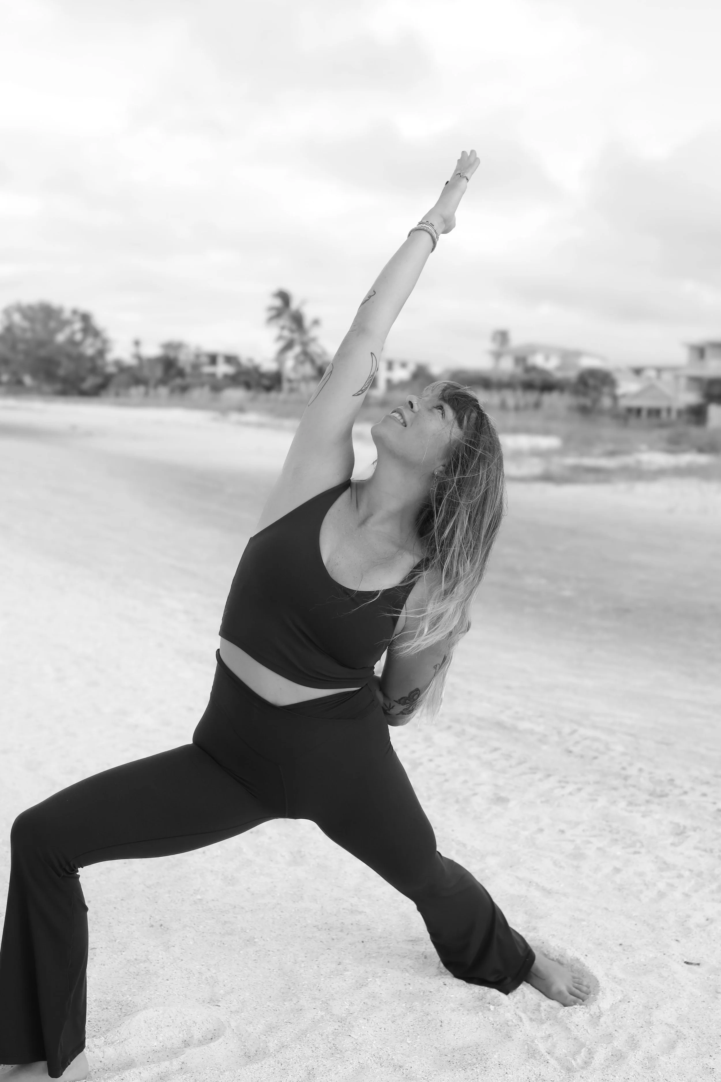 A woman practicing yoga outdoors on a beach, performing a Warrior pose with her arms raised and feet apart.