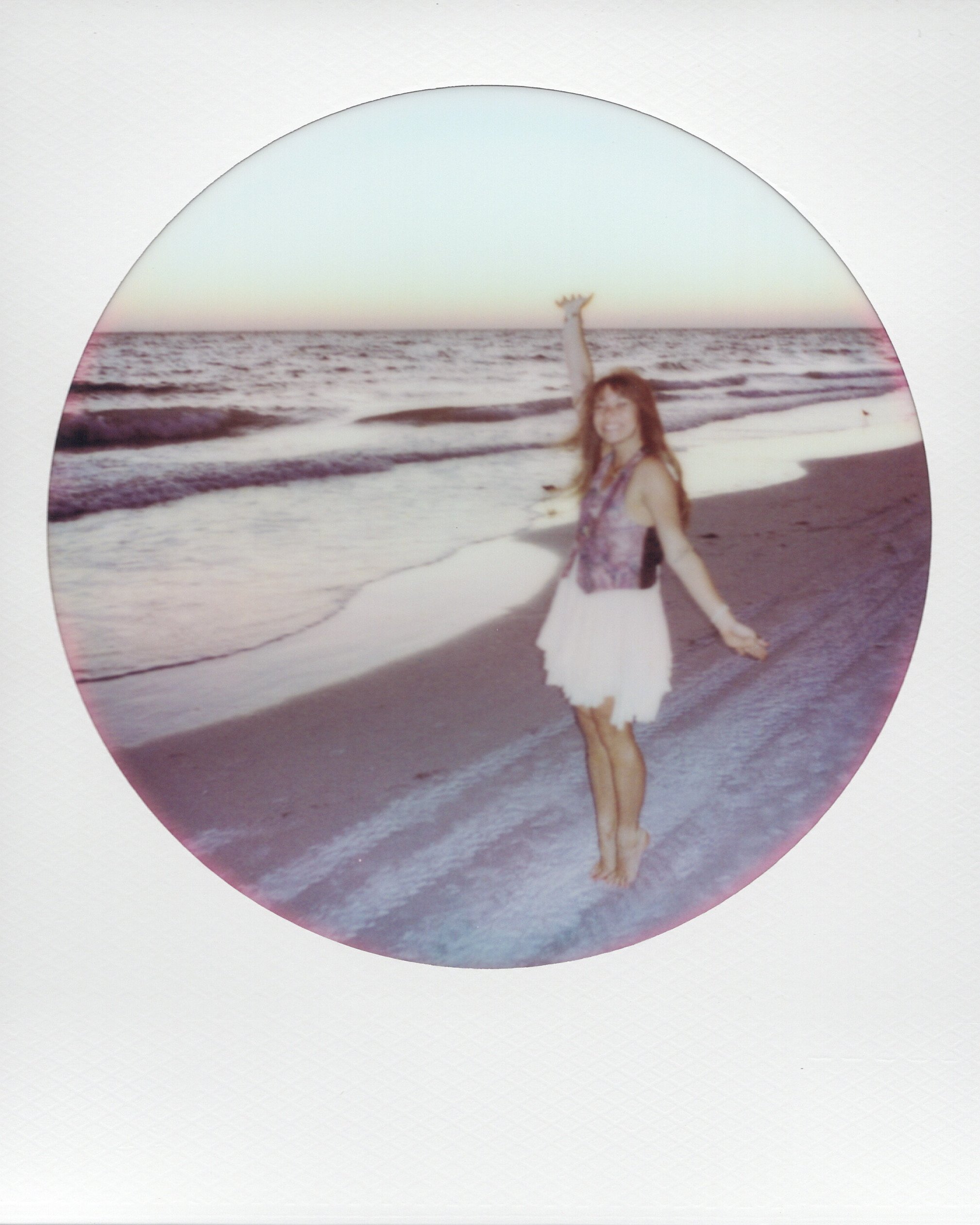 A woman in a white skirt and sleeveless top walks on the beach with her arms outstretched, holding a seashell in her right hand, near the shoreline with small waves, during sunset or sunrise.