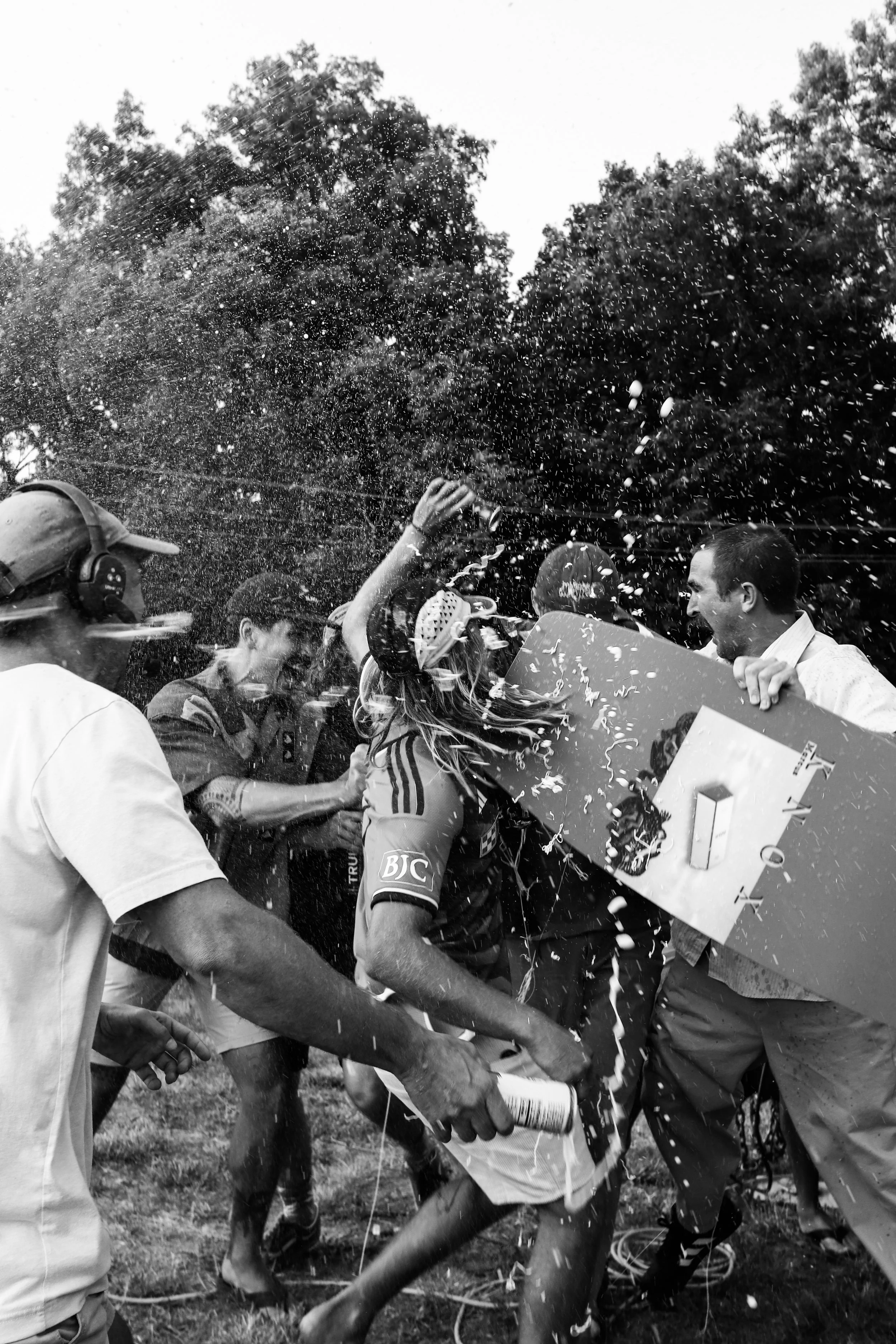 People celebrating outdoors, spraying champagne, and holding a prize check, with trees in the background.