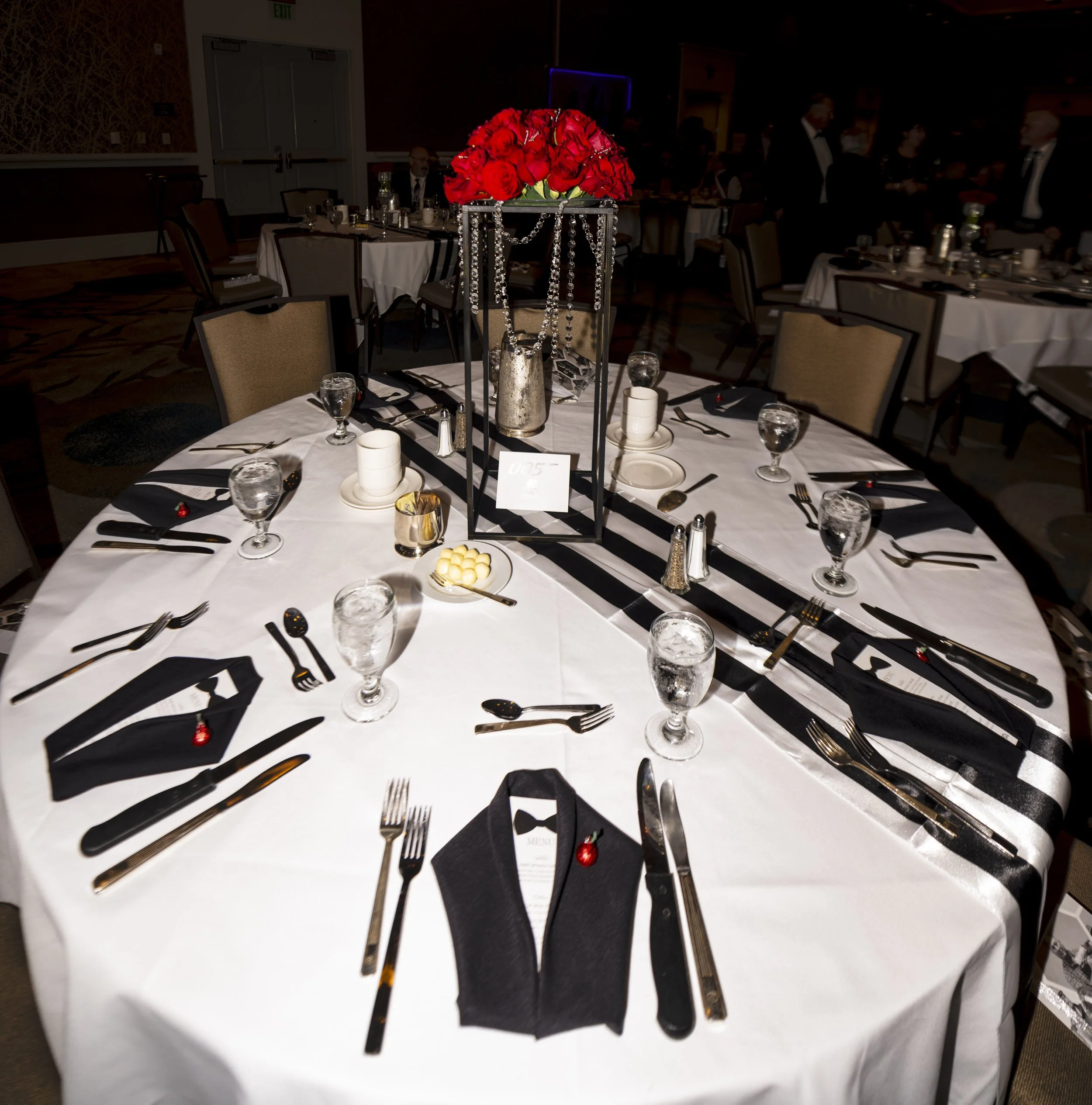 James Bond-themed table scape featuring tuxedo folded napkins, menus with red rose boutonniere chocolates, red rose centerpieces, and crystal accents for a cohesive event design.