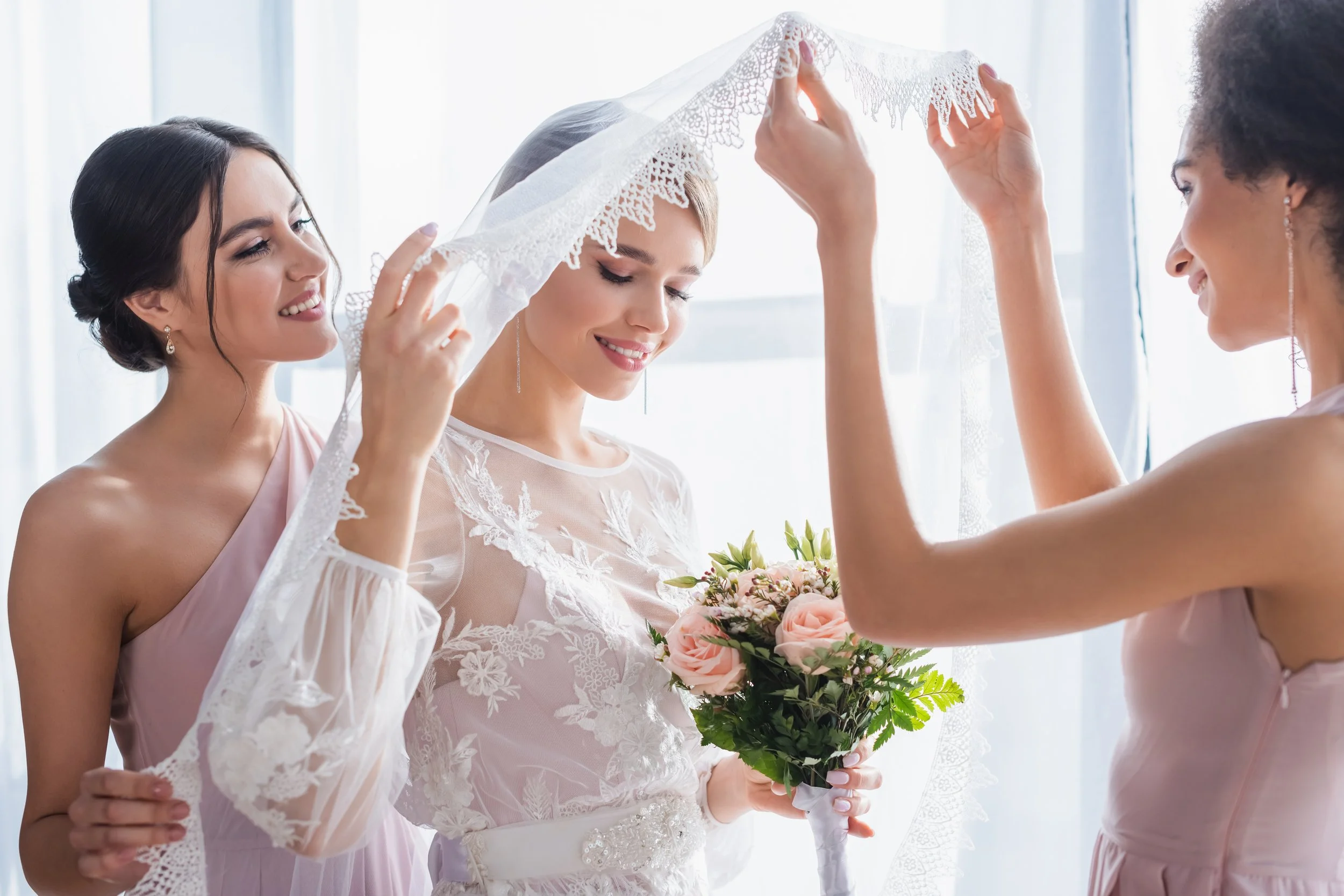 A bride in a lace wedding dress is holding a bouquet of pink roses as two bridesmaids lift a lace veil over her head.